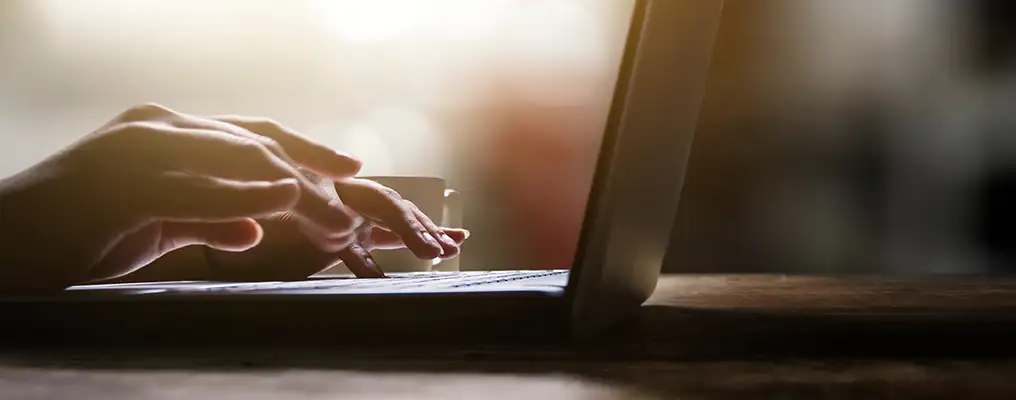 Hands typing on a laptop keyboard at a sunlit wooden desk, with a cup in the background, conveying focus and productivity.