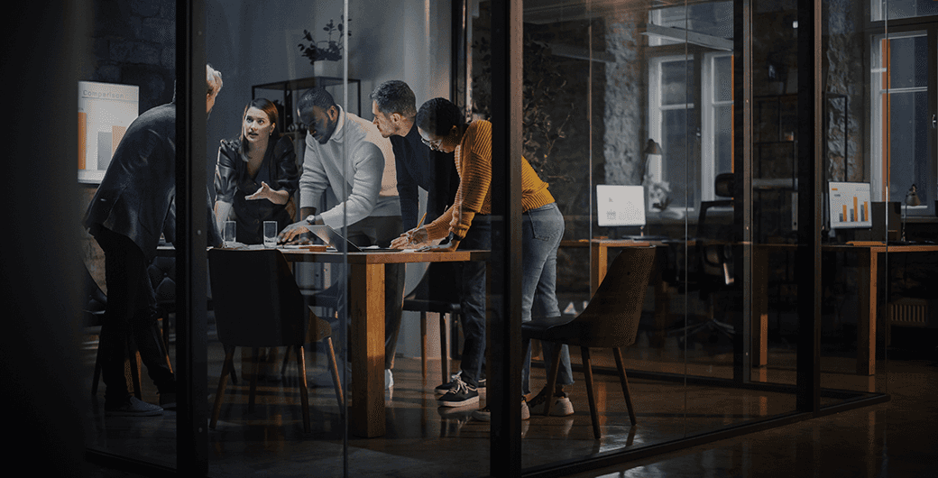 Group of professionals collaborating around a conference table in a modern glass-walled office at night, reviewing documents and laptops.
