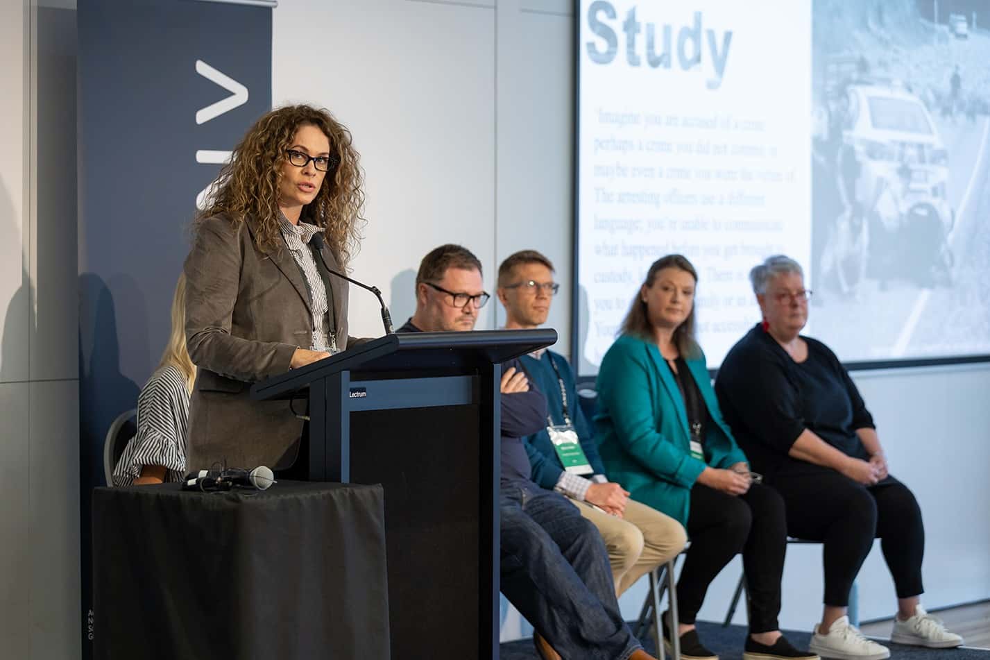 Woman with curly hair and glasses speaking at a podium, four panelists seated behind her in a conference room.