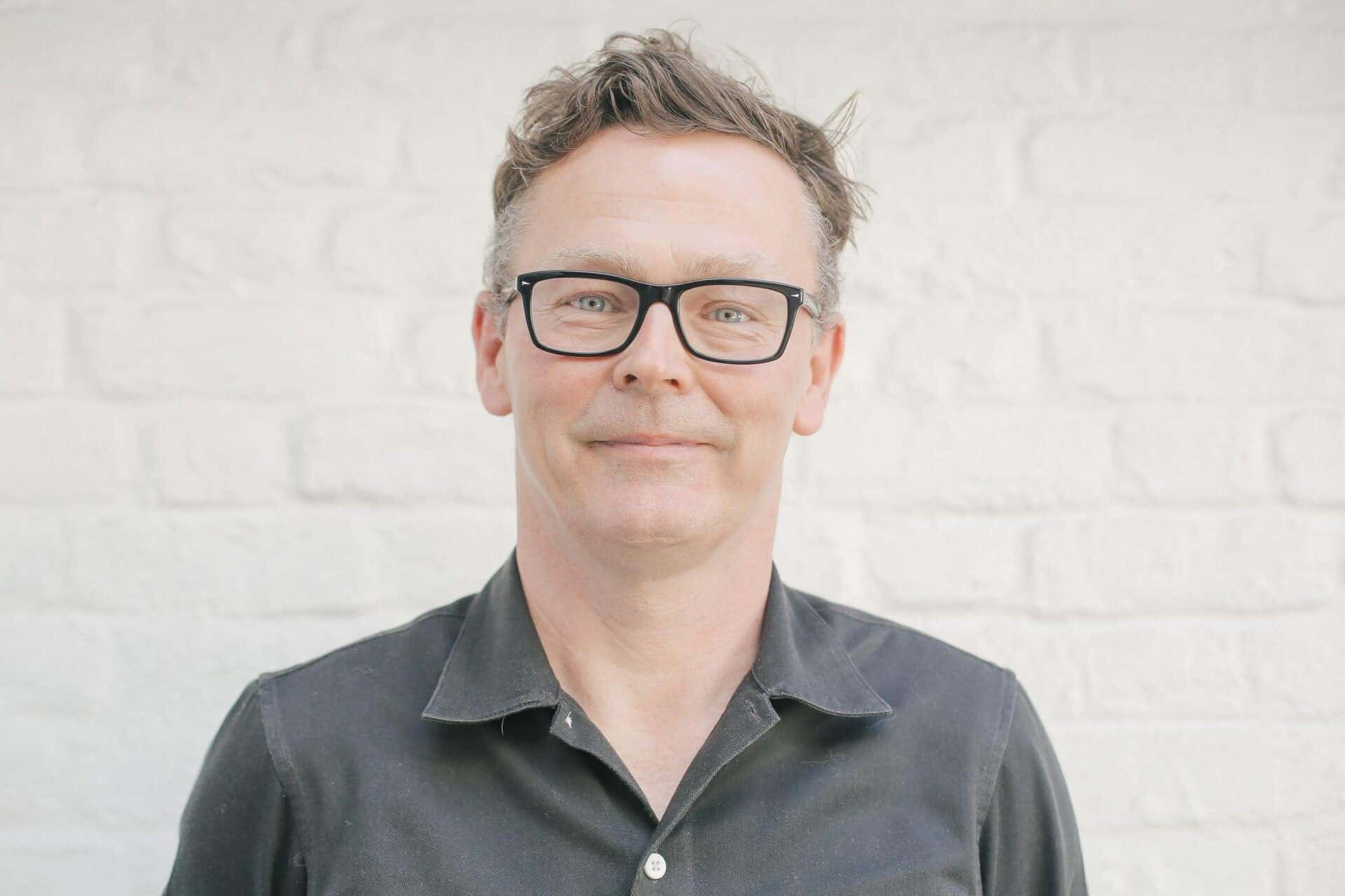 Man with glasses and short, wavy hair wearing a dark shirt, standing against a white brick wall.