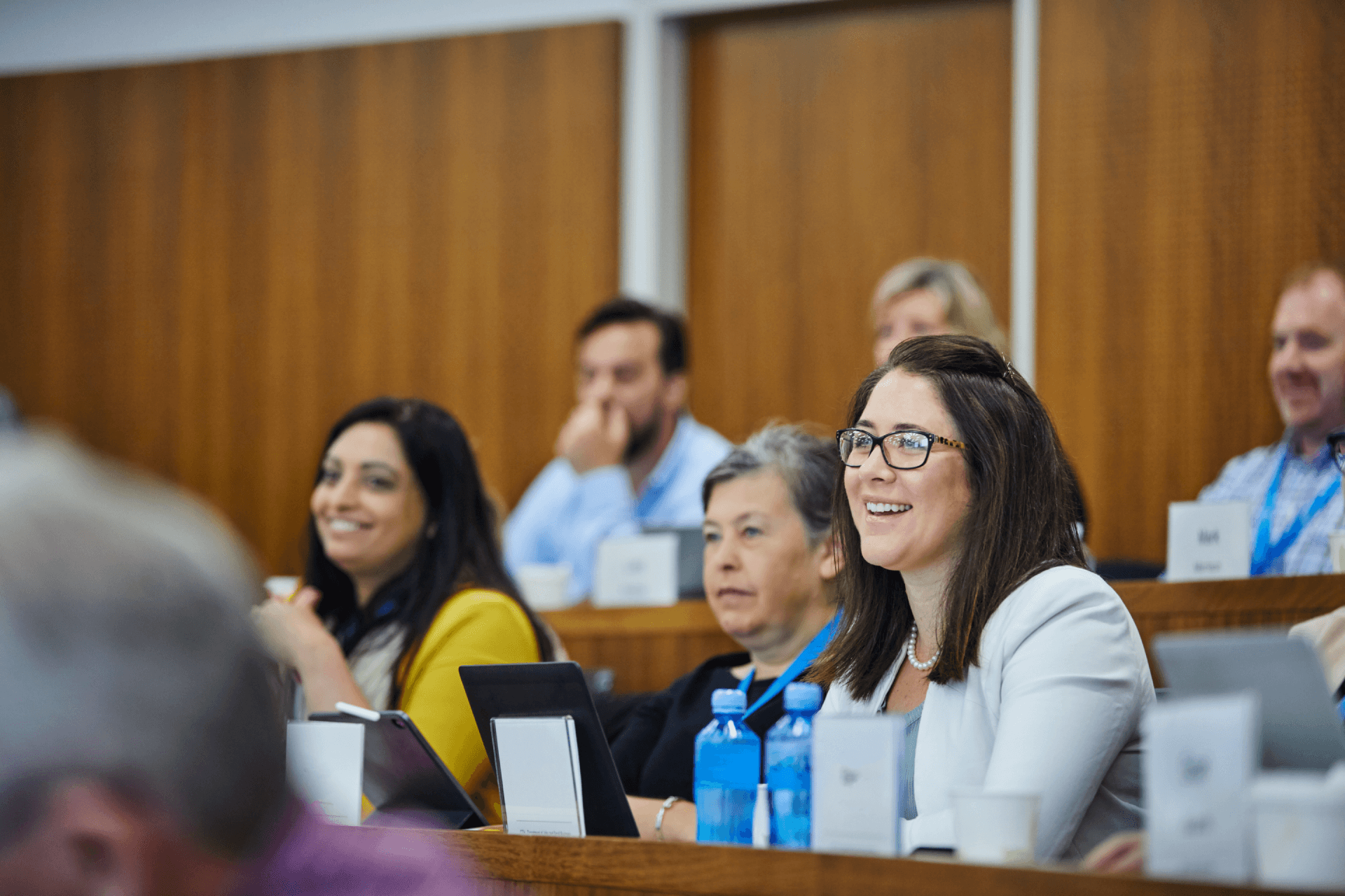 Smiling people sitting in a conference room, engaged and attentive, with laptops, tablets, and notebooks in front of them.