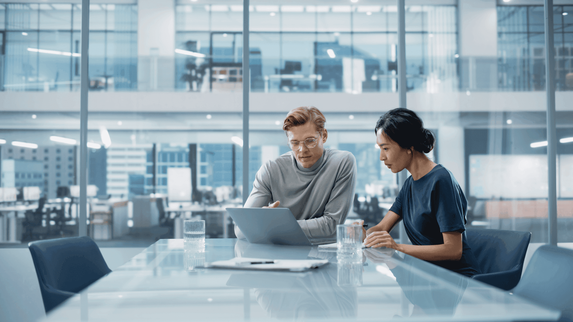 Two people collaborate at a conference table with a laptop, in a modern office with glass walls and a city view.