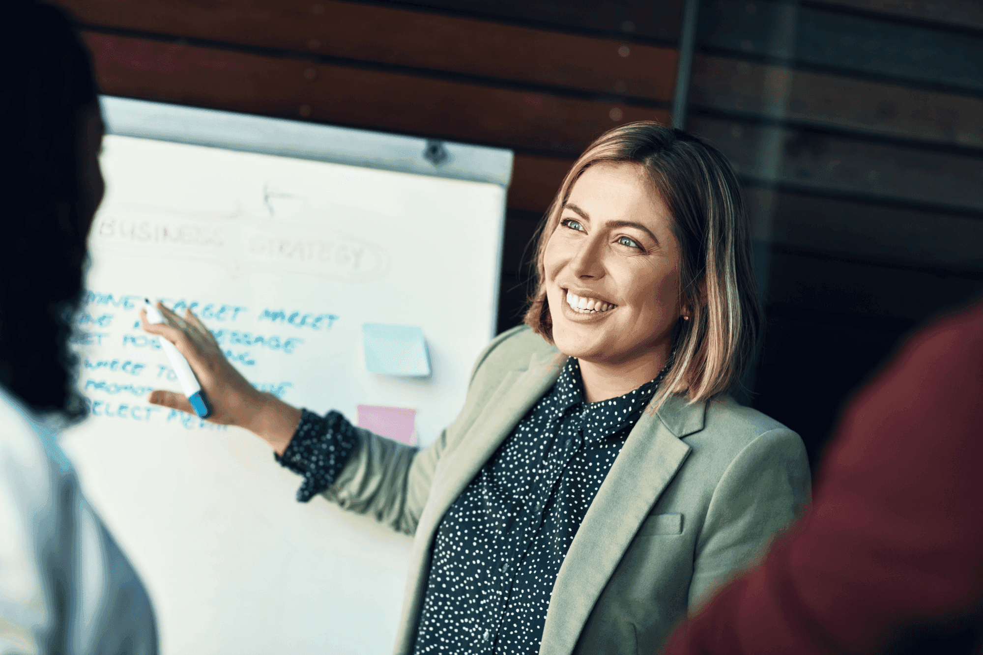 A woman in a green blazer smiles while presenting a business strategy on a whiteboard during a meeting.