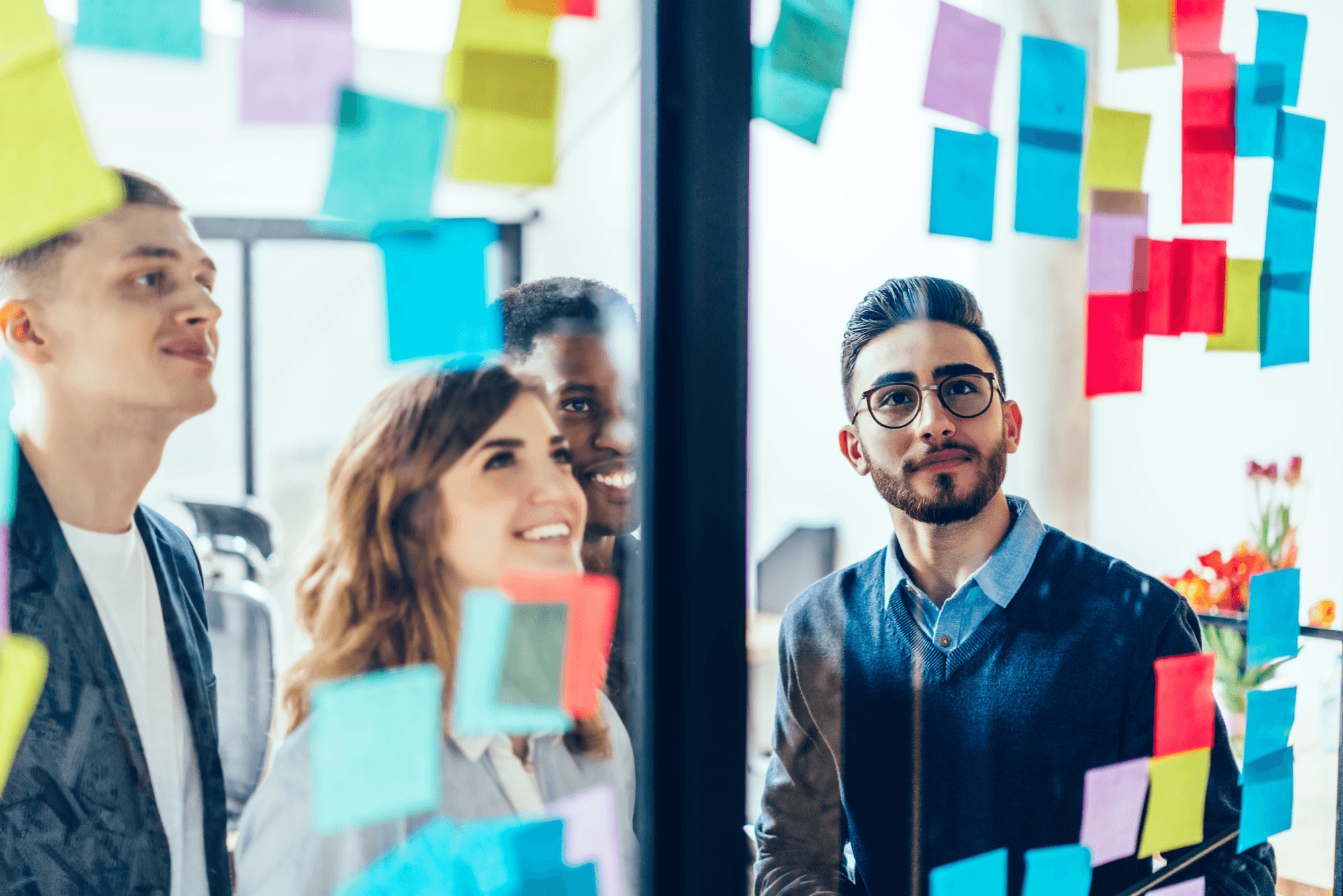 A group of four people stands in front of a glass wall, covered with colorful sticky notes, discussing ideas in a bright office setting.