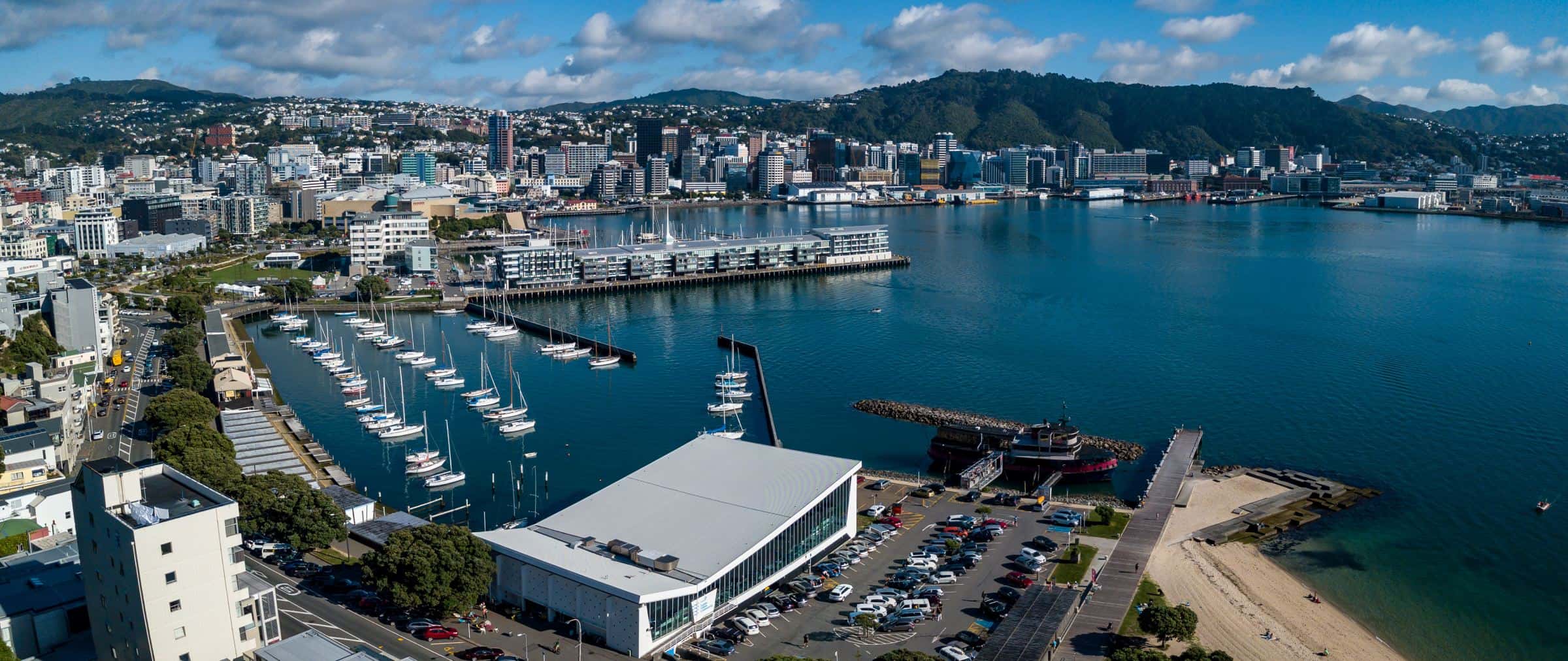 Aerial view of a coastal city with a marina, high-rise buildings, lush hills, and a sandy beach under a blue sky with scattered clouds.