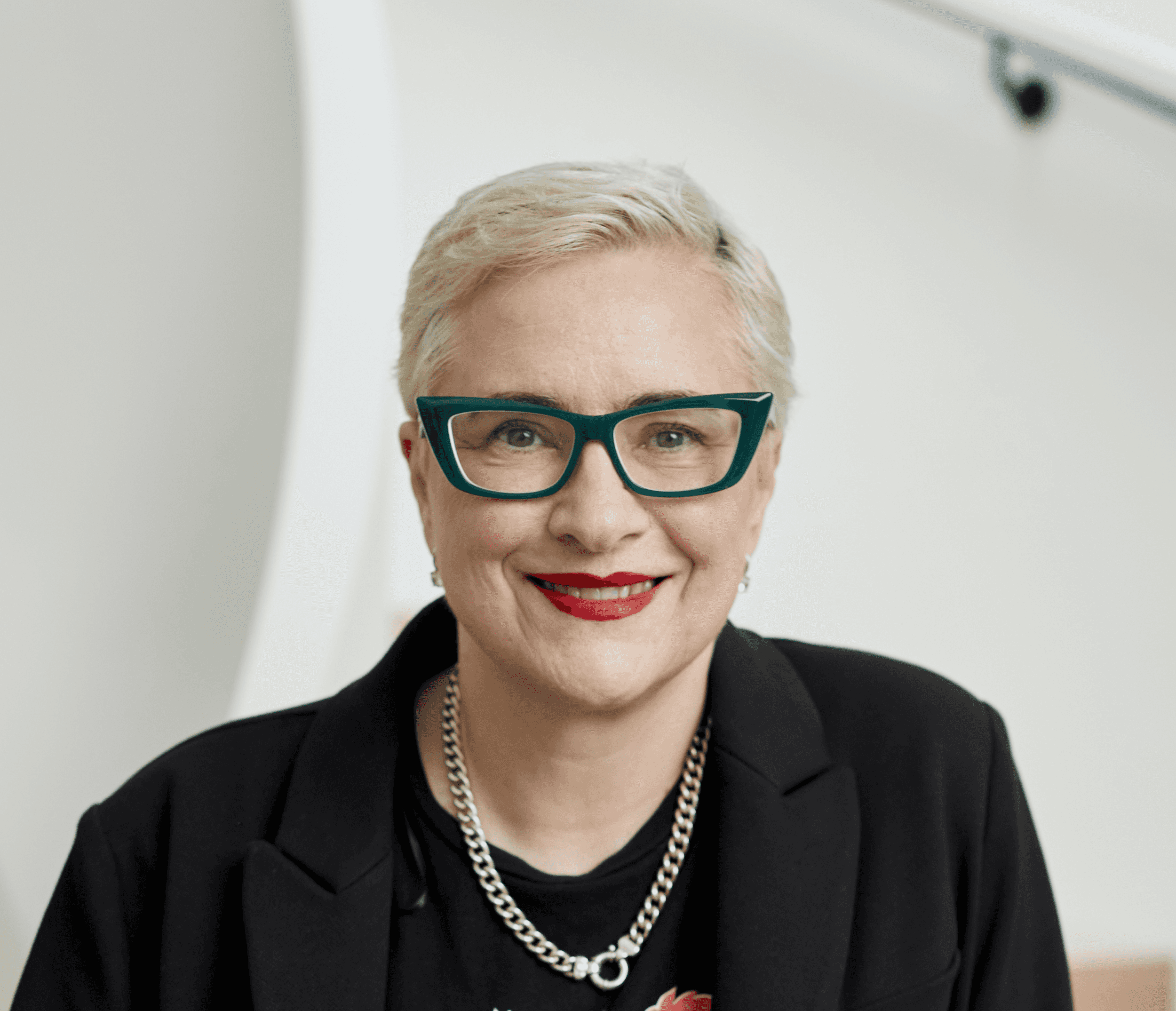 Person with short blond hair, wearing teal glasses, a black blazer, a chain necklace, and red lipstick, smiling at the camera.