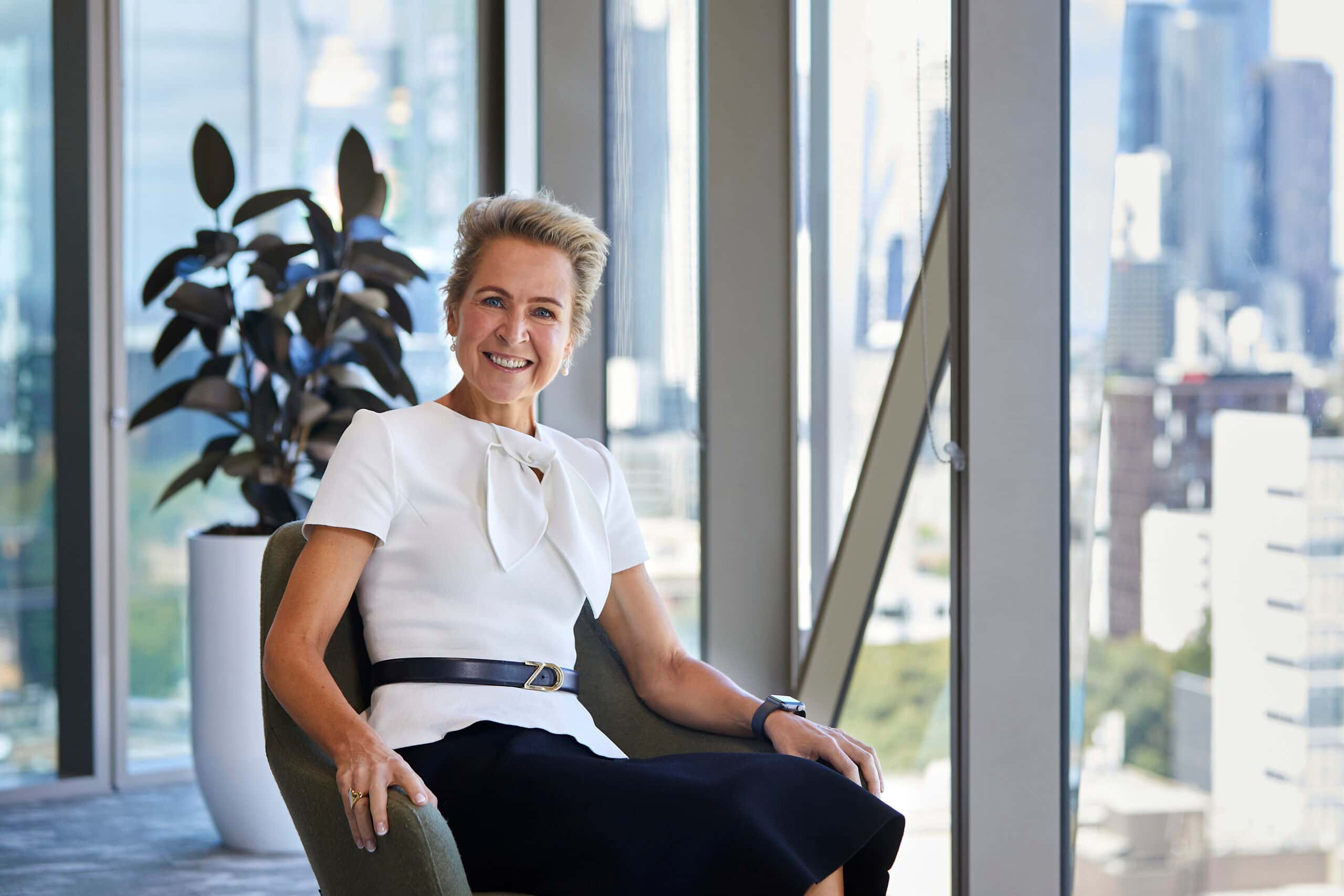 Smiling professional woman in white blouse and black skirt seated in office chair by floor-to-ceiling windows with city view.