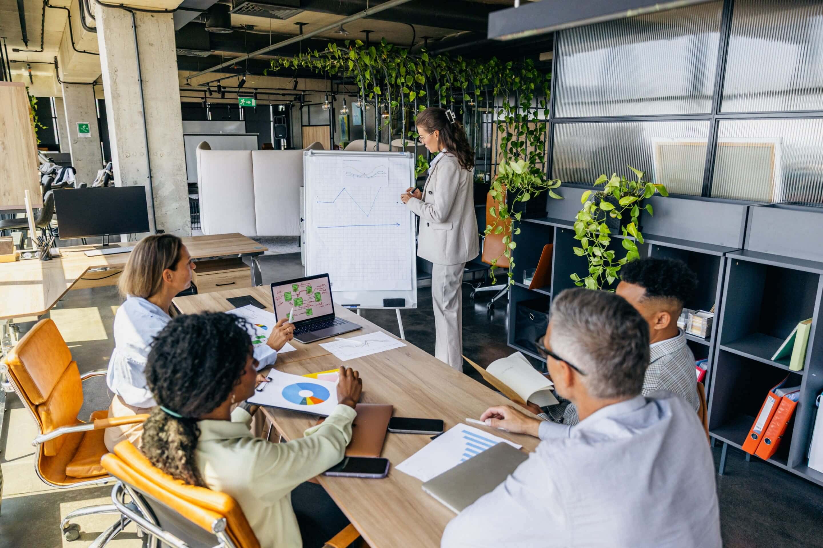 A person presents graphs on a flipchart to four colleagues in a modern office with plants and laptops on the table.