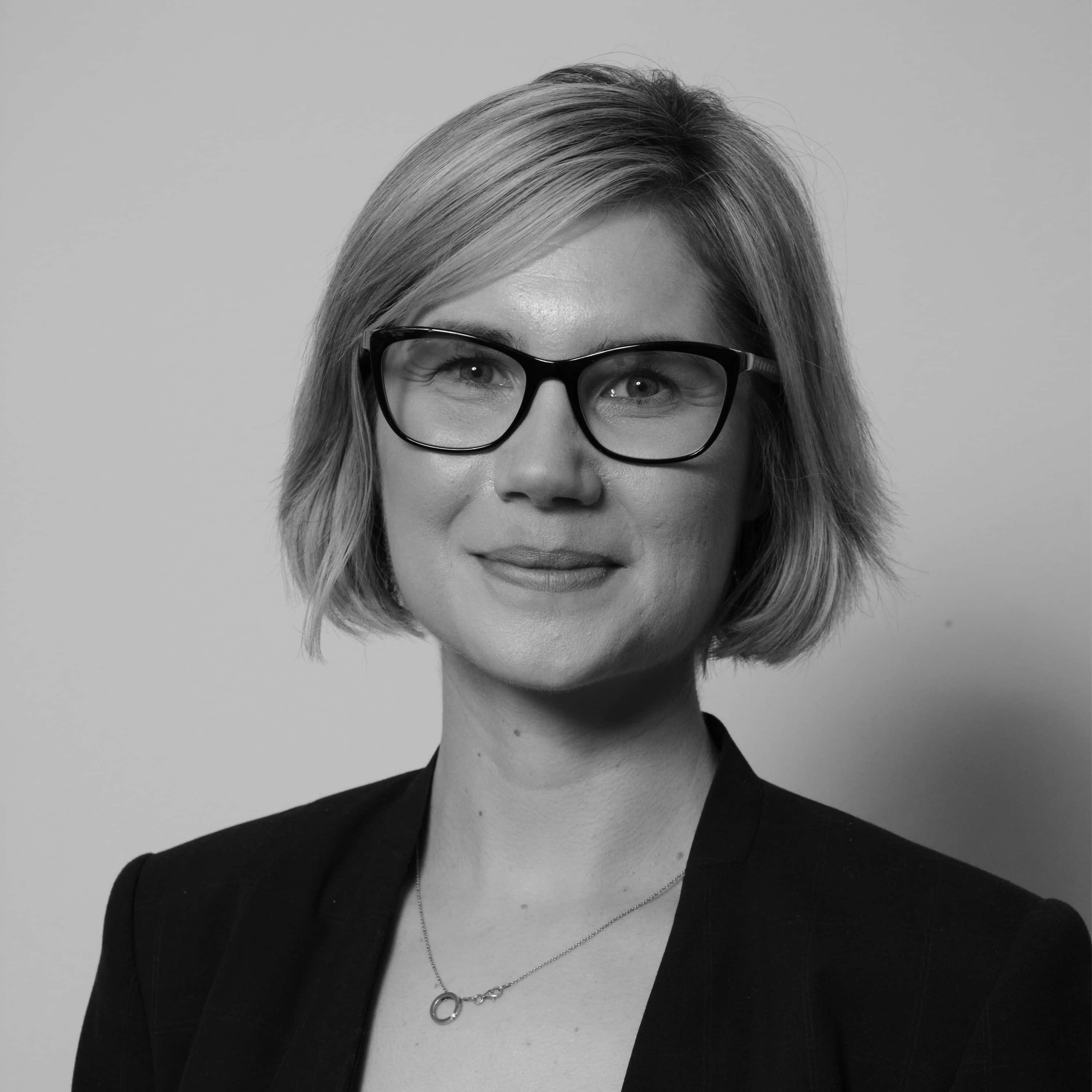 Black and white professional headshot of a woman with short blonde hair, glasses, blazer, and a delicate necklace, smiling softly.
