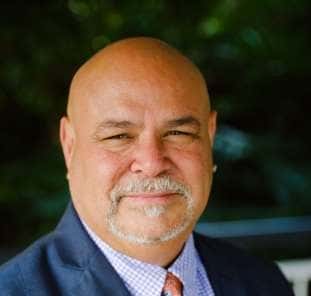 Bald man with a mustache and goatee, wearing a suit and tie, smiling slightly, set against a blurred outdoor background.