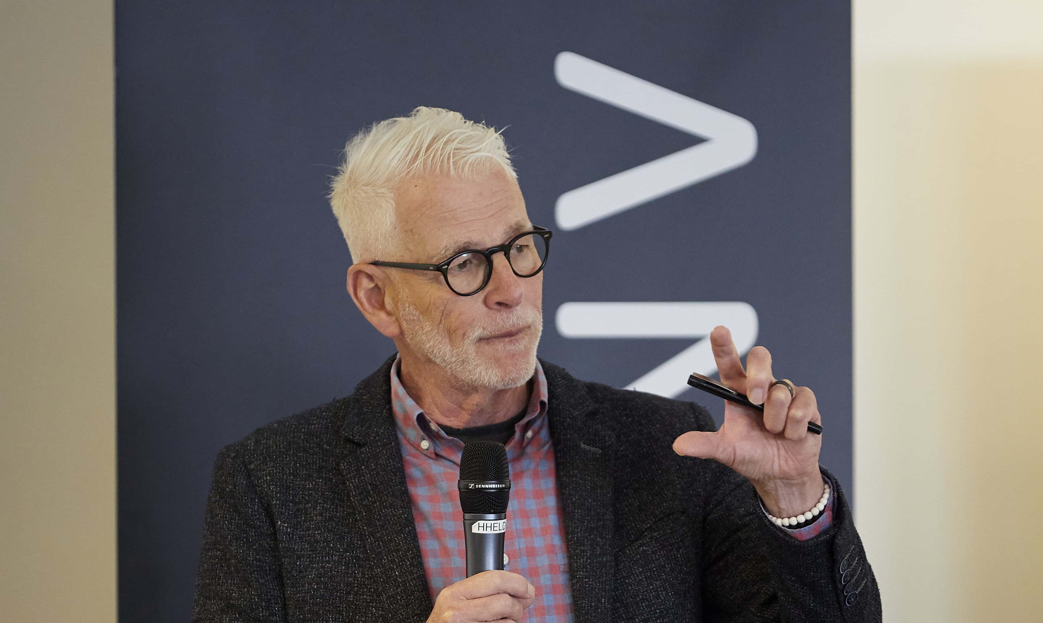 Elderly man with glasses speaking into a microphone, gesturing with a pen. Background features dark banner with white emblem.