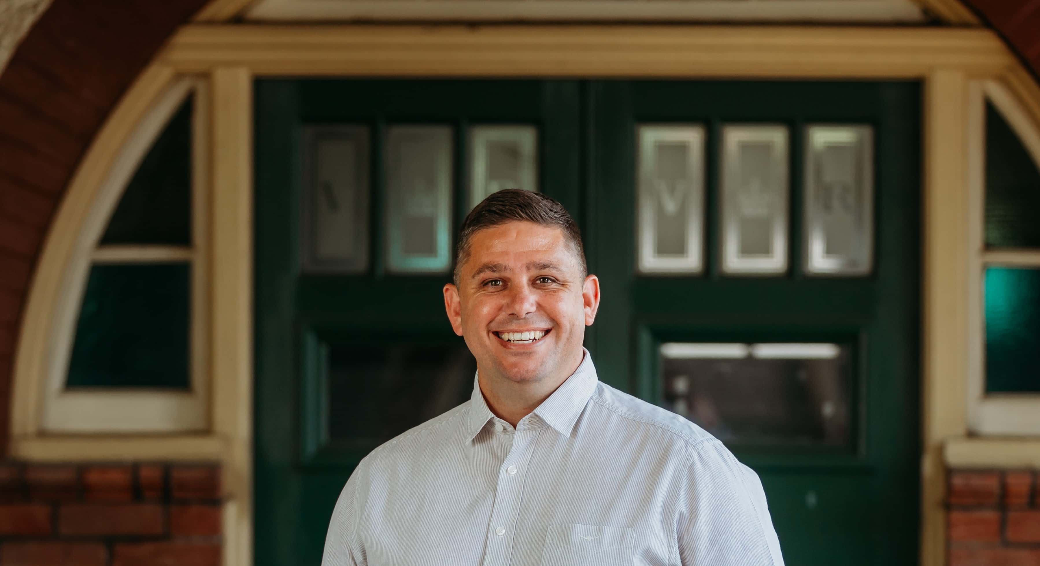 Smiling man in a light shirt standing in front of a green door with decorative glass panels.