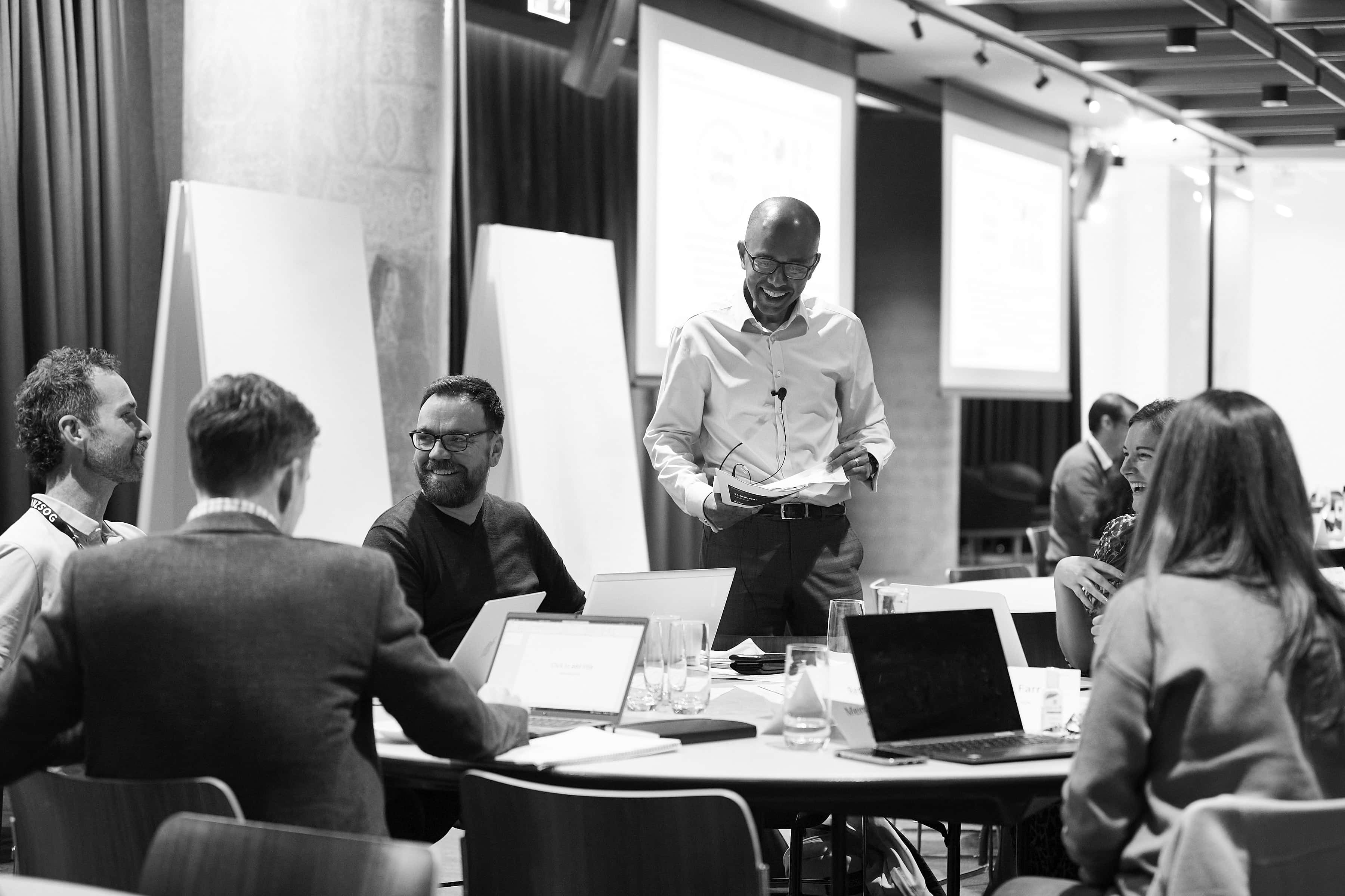 Smiling presenter stands with papers while colleagues around a table laugh and work on laptops in a conference room.