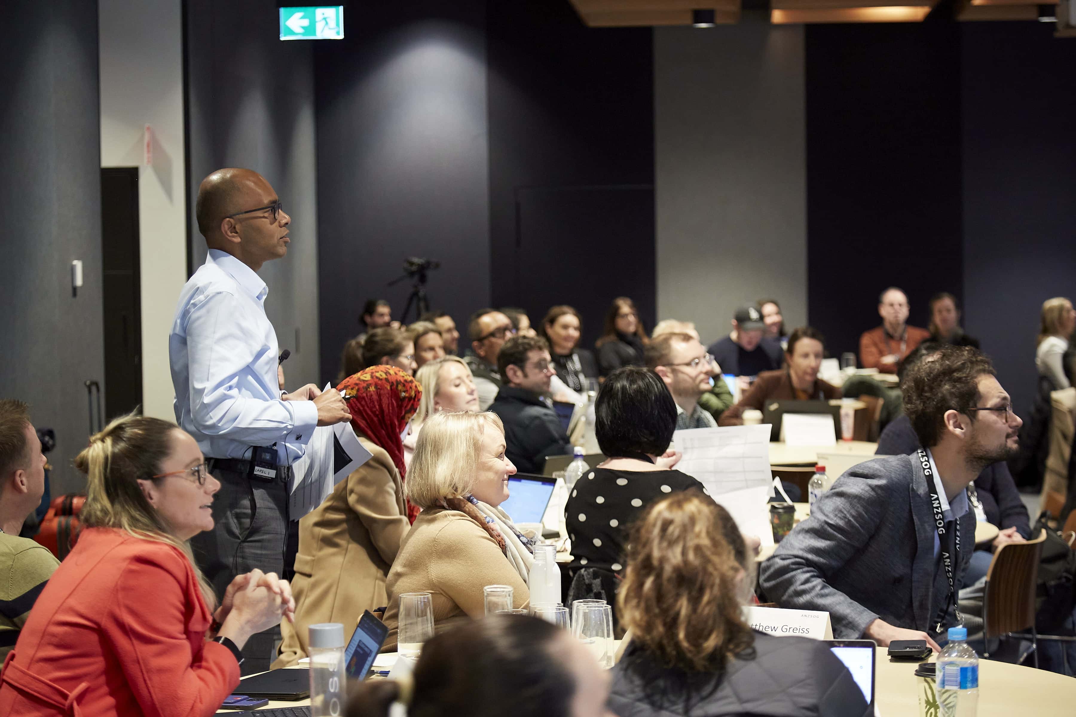 Attendees at a conference seated at tables with laptops listen as a man stands up and speaks to the room.