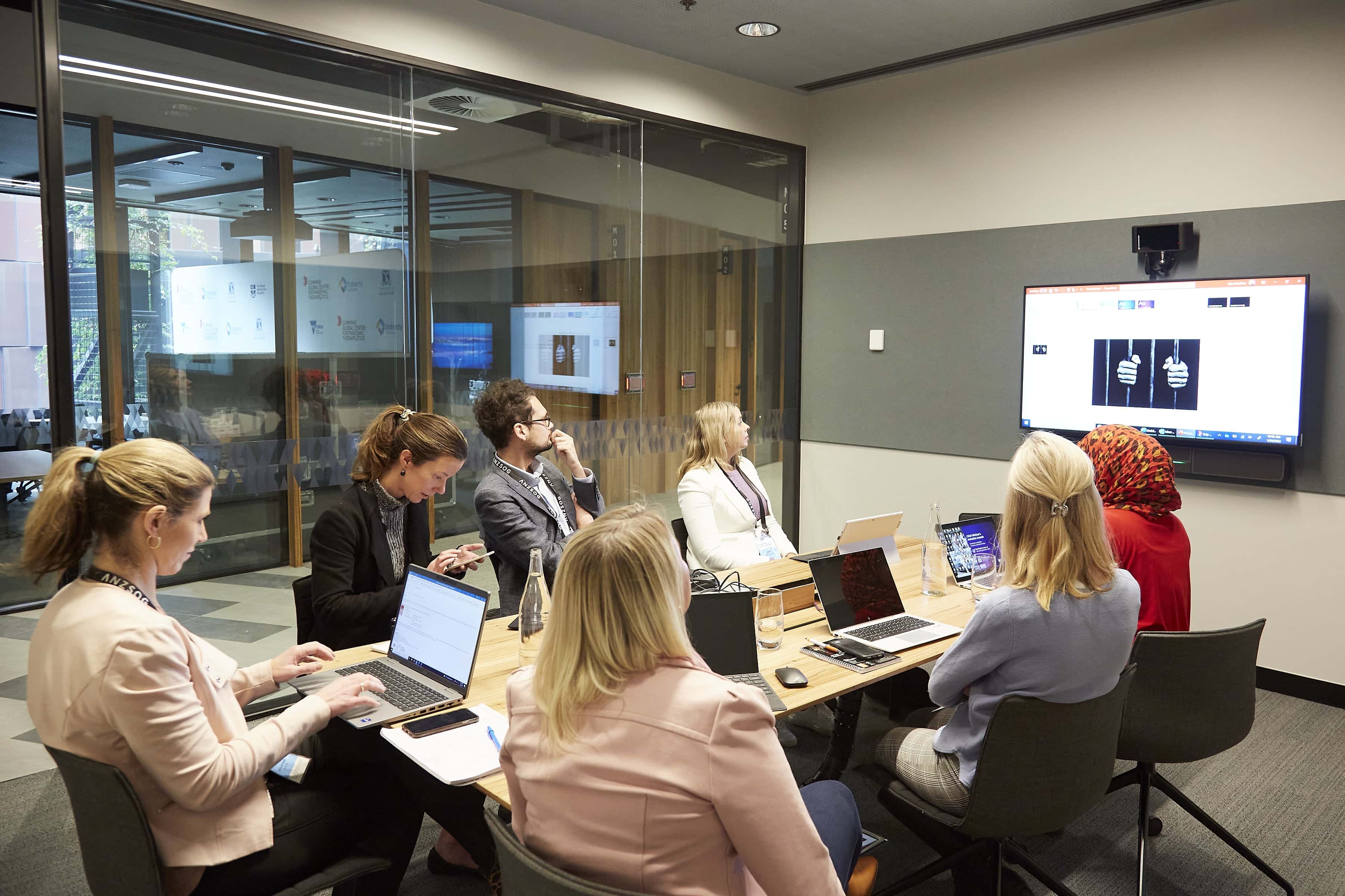 Seven professionals in a glass-walled conference room seated around a table, viewing a presentation on a wall-mounted monitor.