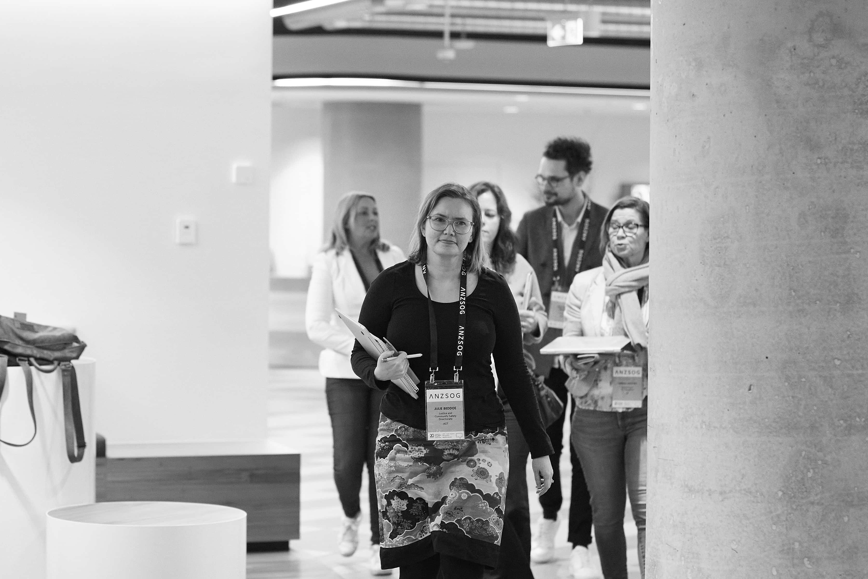 Black-and-white photo of a woman wearing a conference badge leading a small group indoors, holding a clipboard and pen.