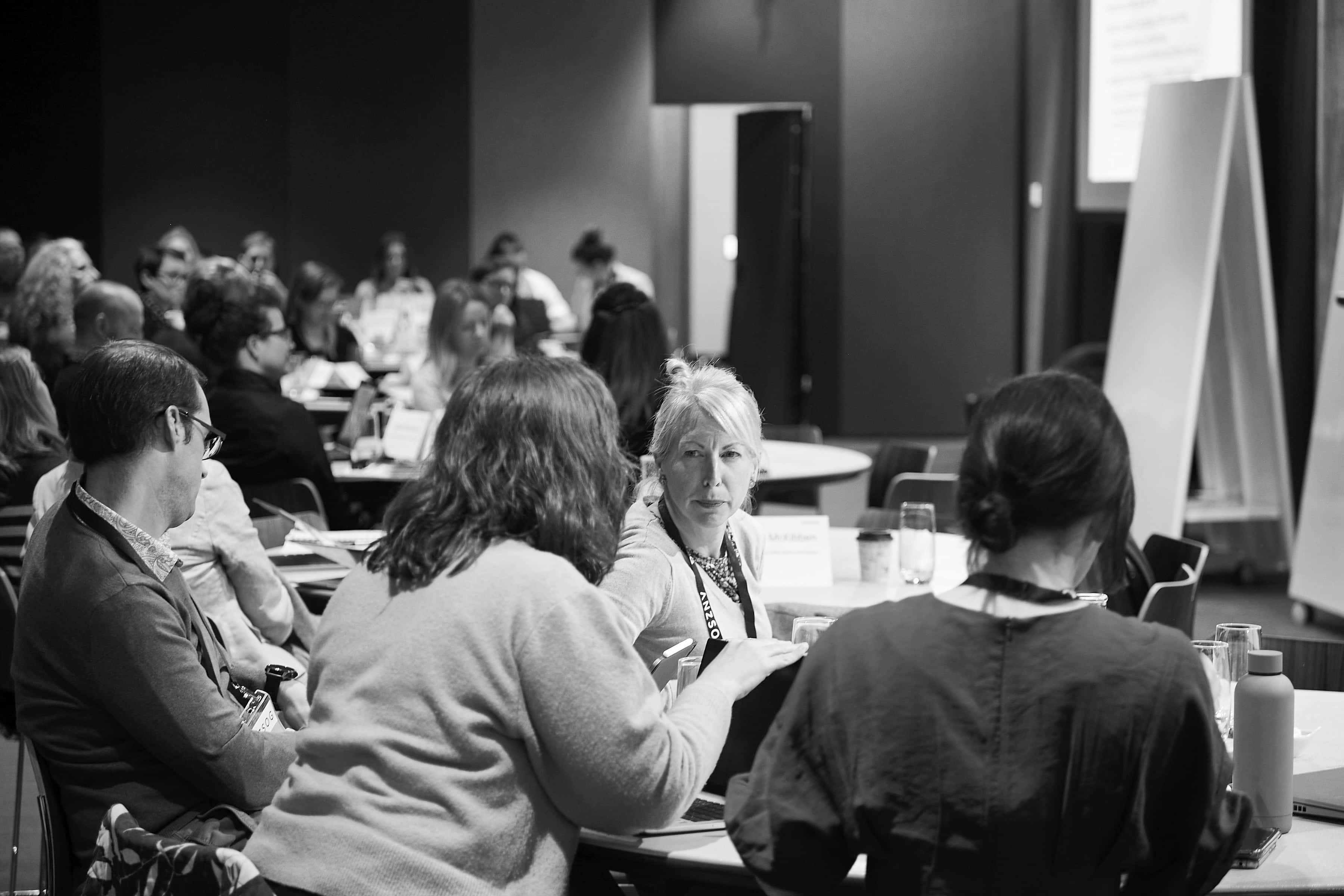 Black-and-white photo of a workshop: a light-haired woman looks intently across a table while colleagues talk and take notes.