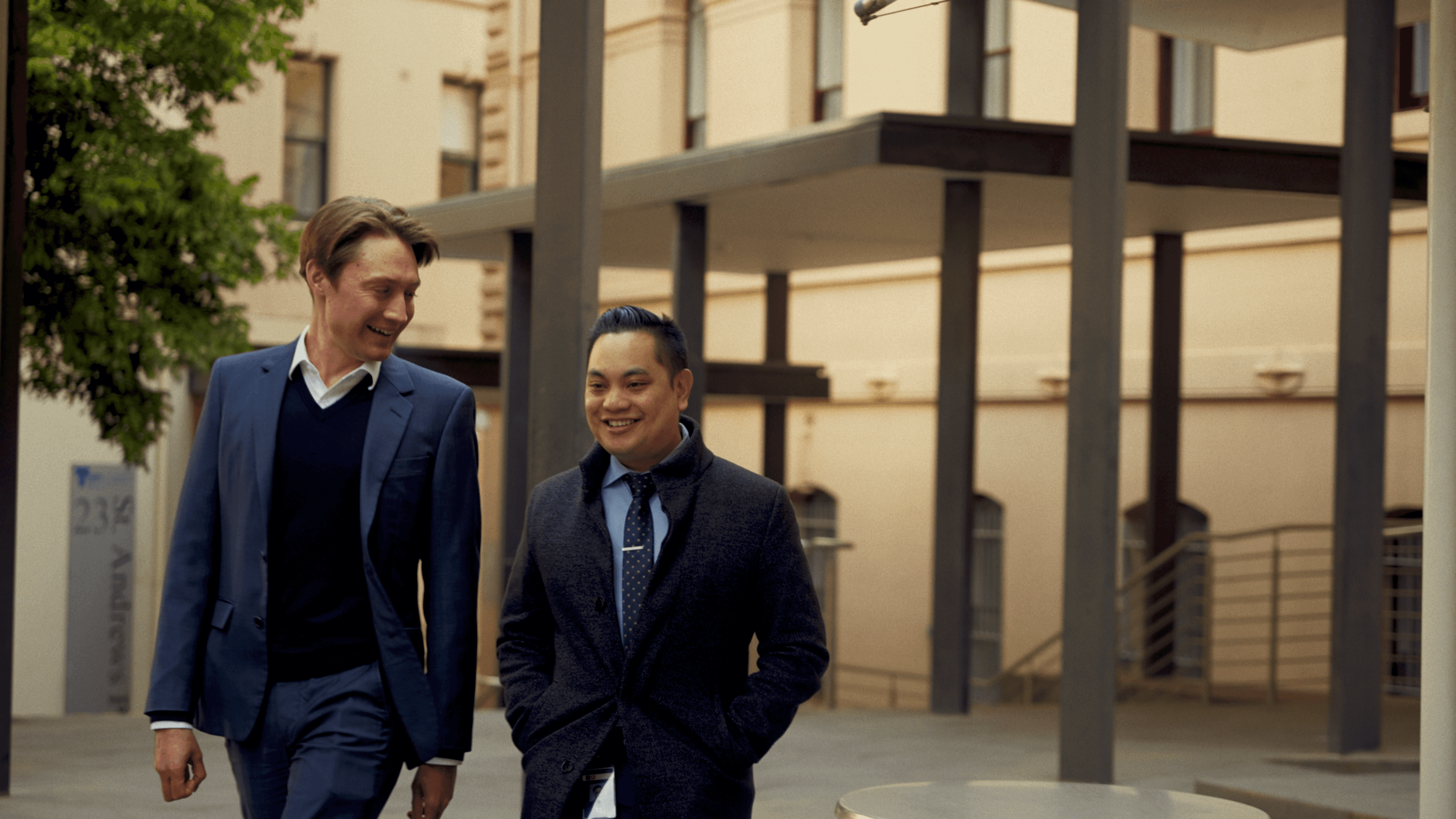 Two men in business attire walk and smile in a modern urban setting with a beige building and pillars in the background.