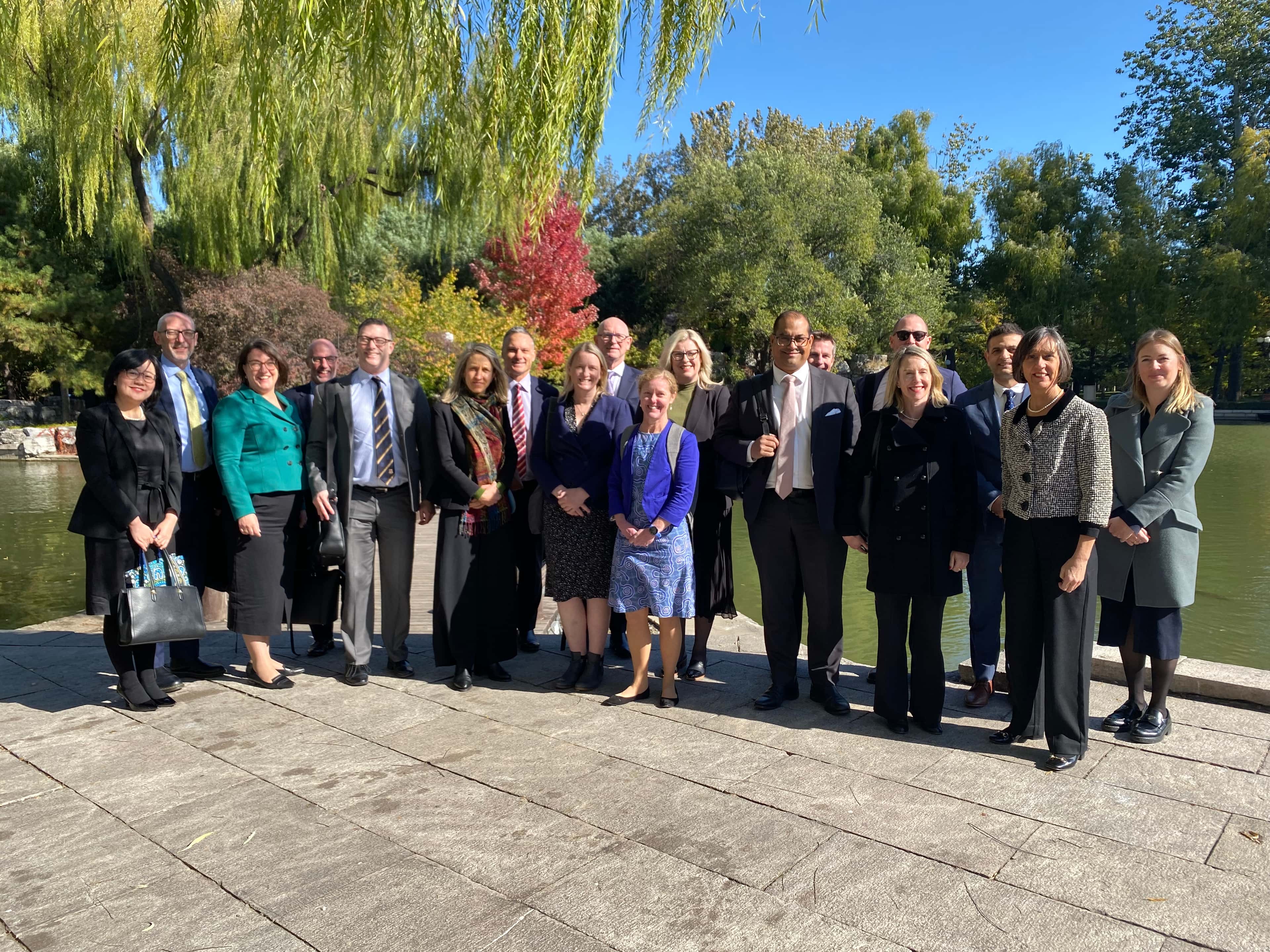 A group of 18 people in formal attire stand outdoors on a sunny day by a lake, with trees and colorful foliage in the background.