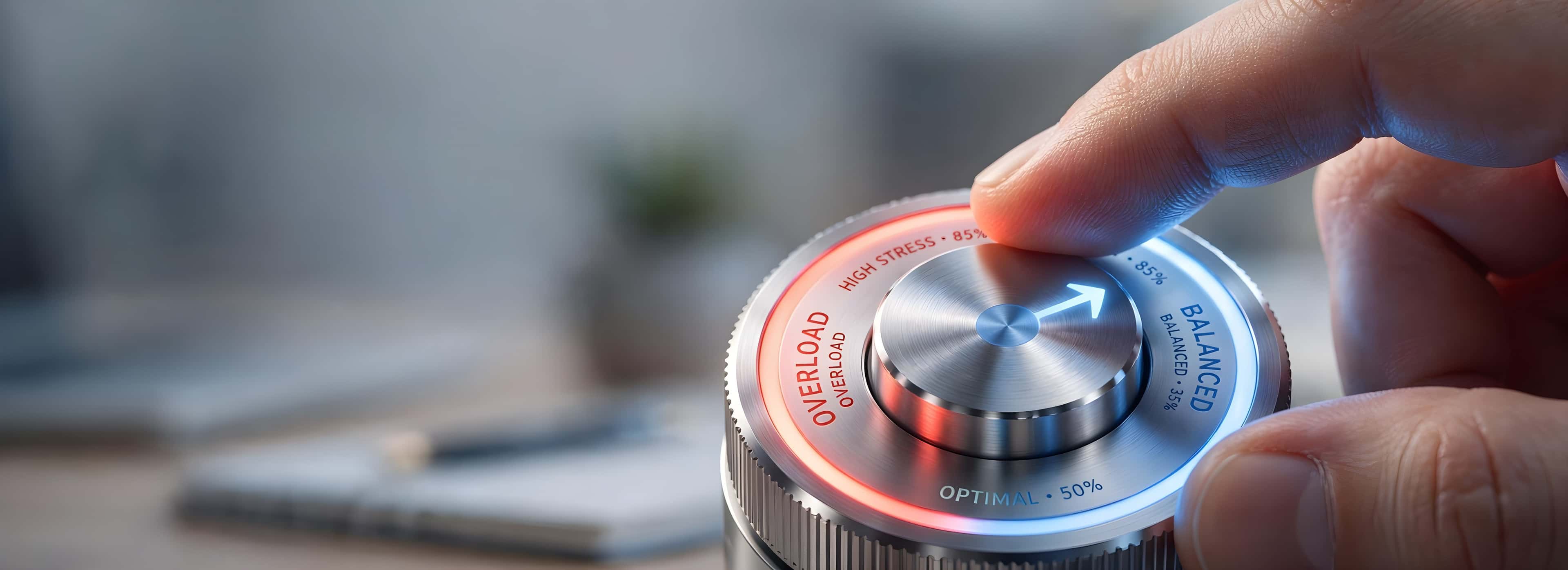 A finger turning a metallic dial labeled from balanced (50%) to overloaded, with a blue and red LED indicator. Background is blurred.