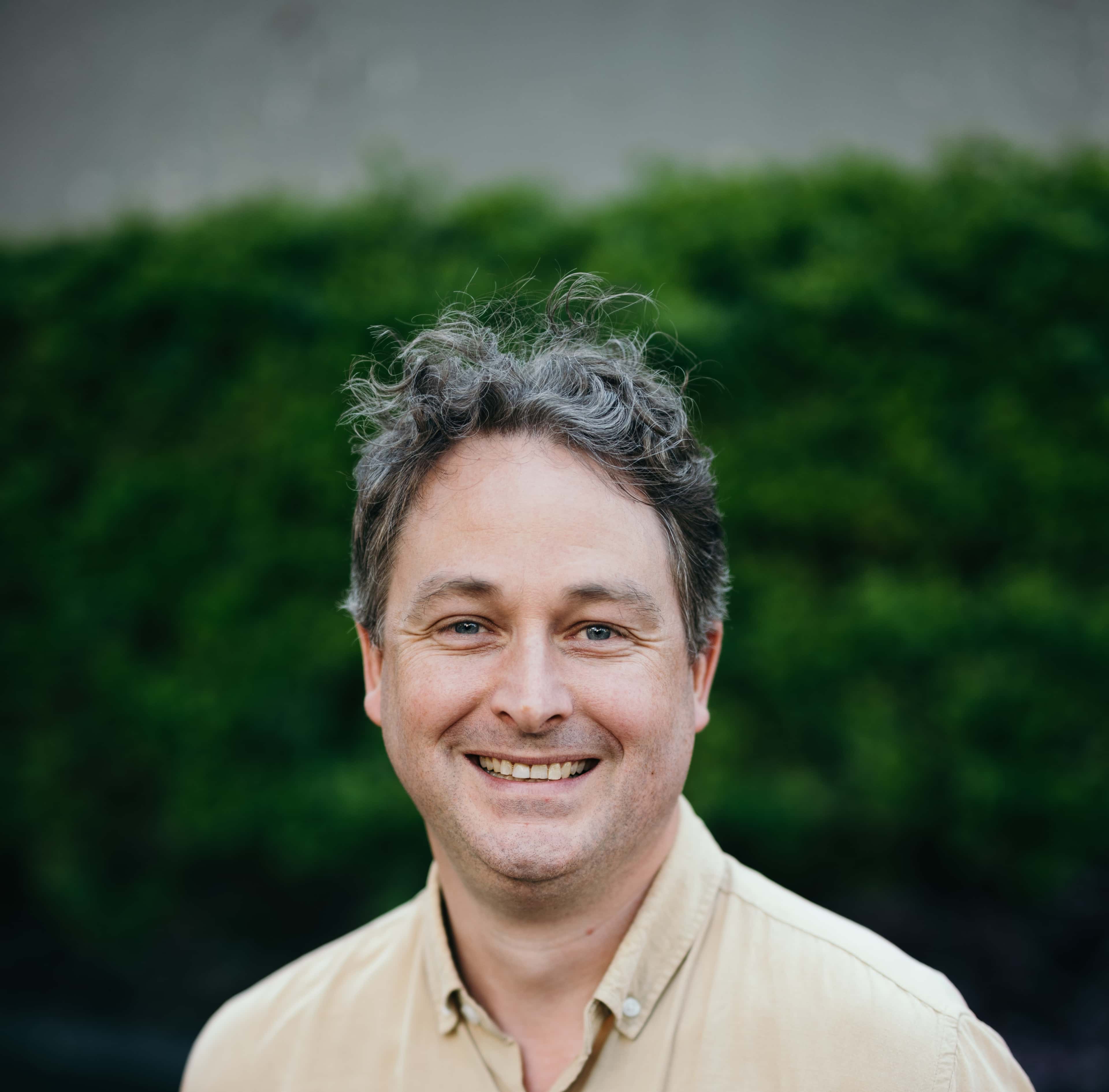 Smiling man with curly hair stands outdoors against a blurred green background, wearing a light-colored shirt.