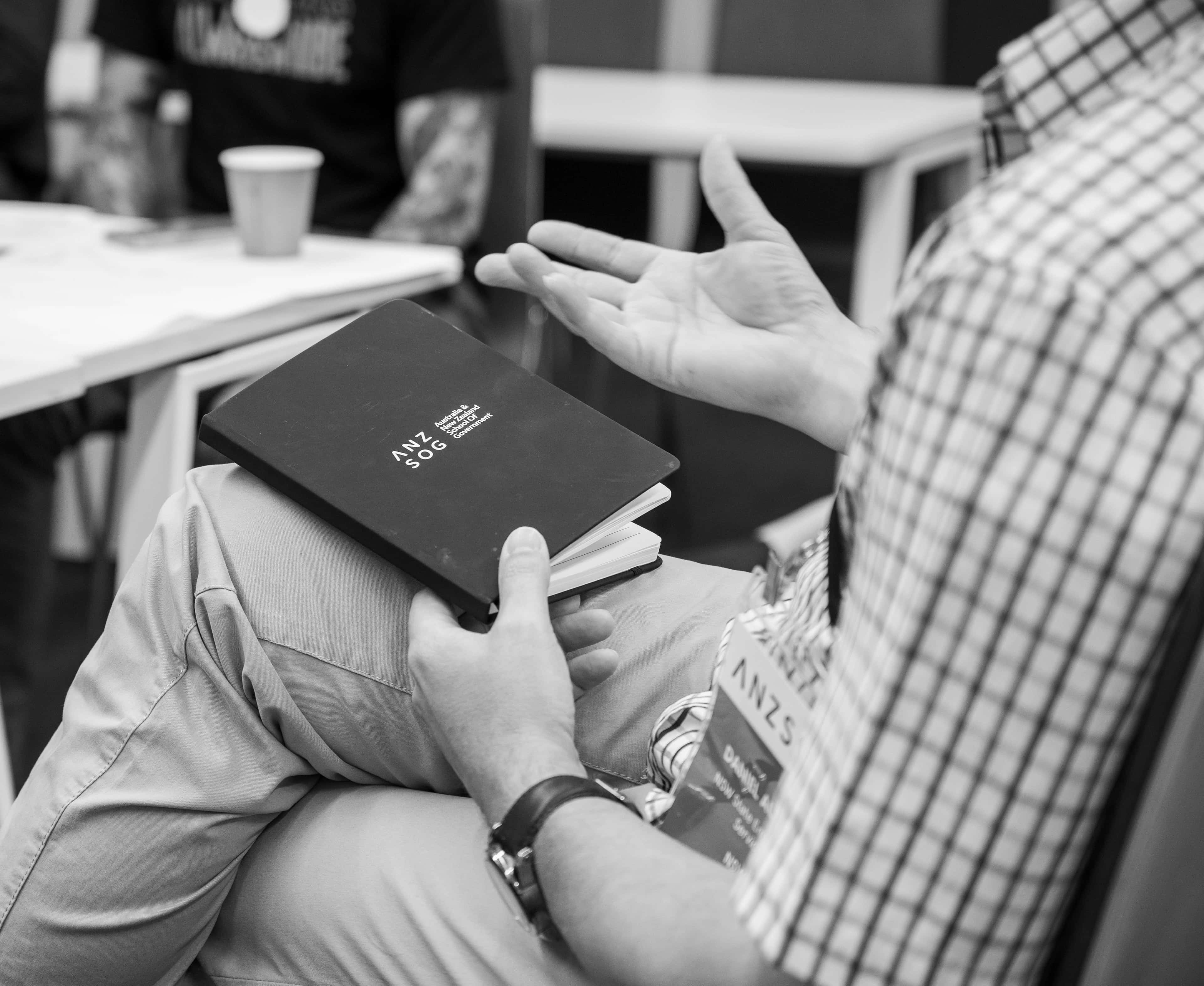 Black-and-white photo of a seated person holding a black notebook labeled "ANZSOG" on their lap, gesturing with one hand.