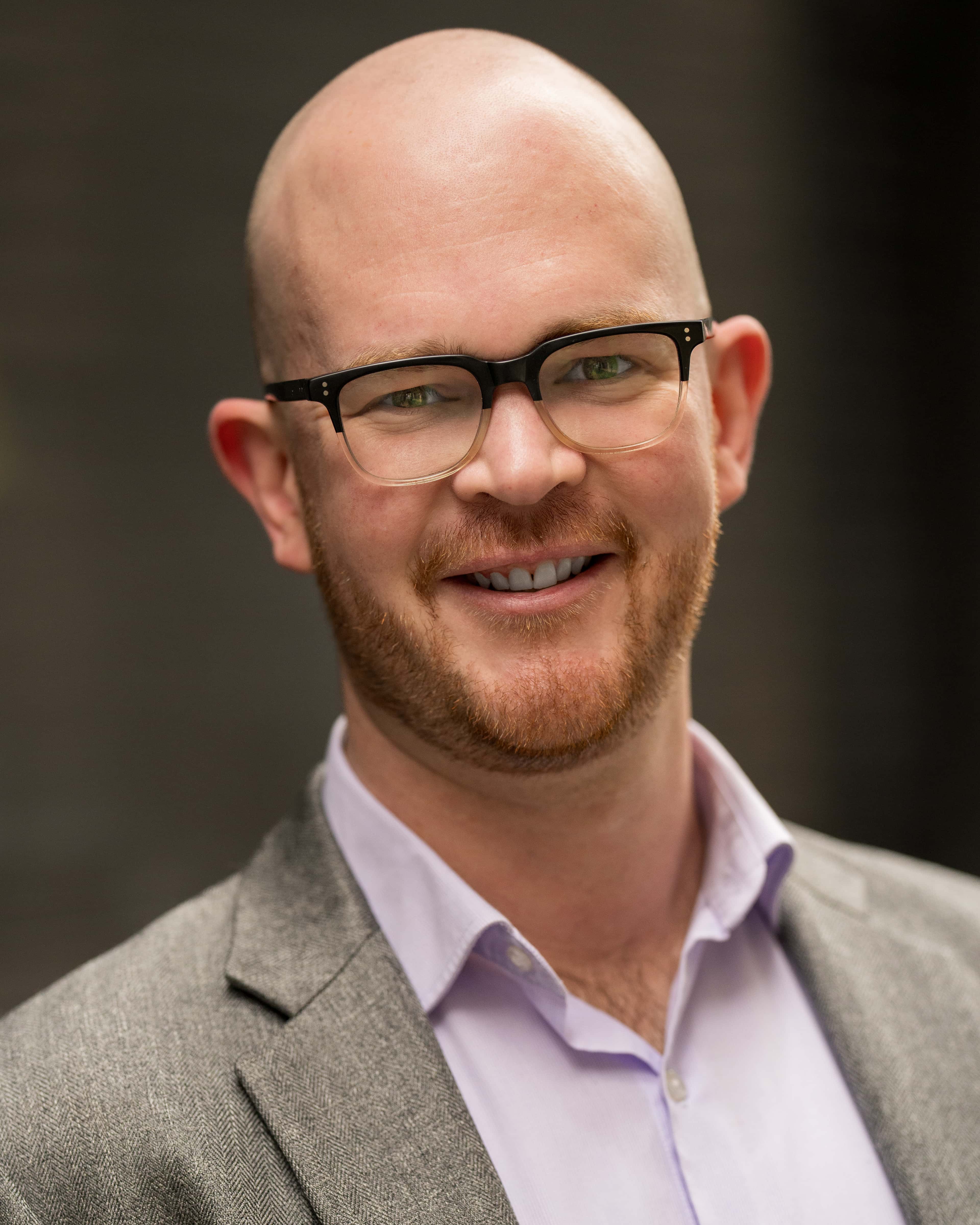 Smiling individual with a bald head and glasses, wearing a light purple shirt and a gray blazer, in front of a muted background.