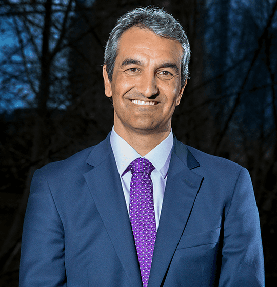 Smiling man in a blue suit and purple tie stands outdoors with trees and a dusk sky in the background.