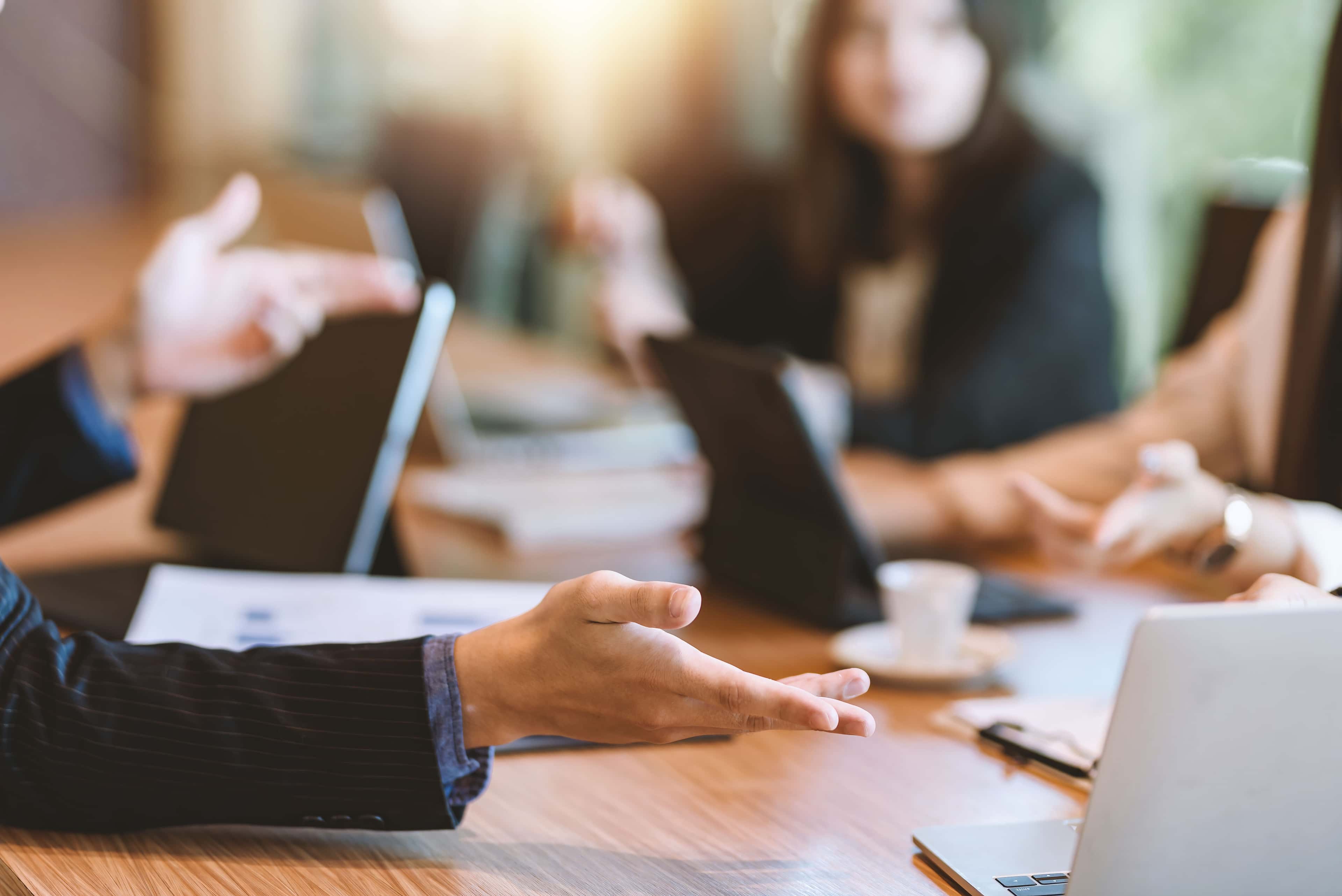 A group of people having a meeting around a table with laptops and documents, one person gesturing with their hand.