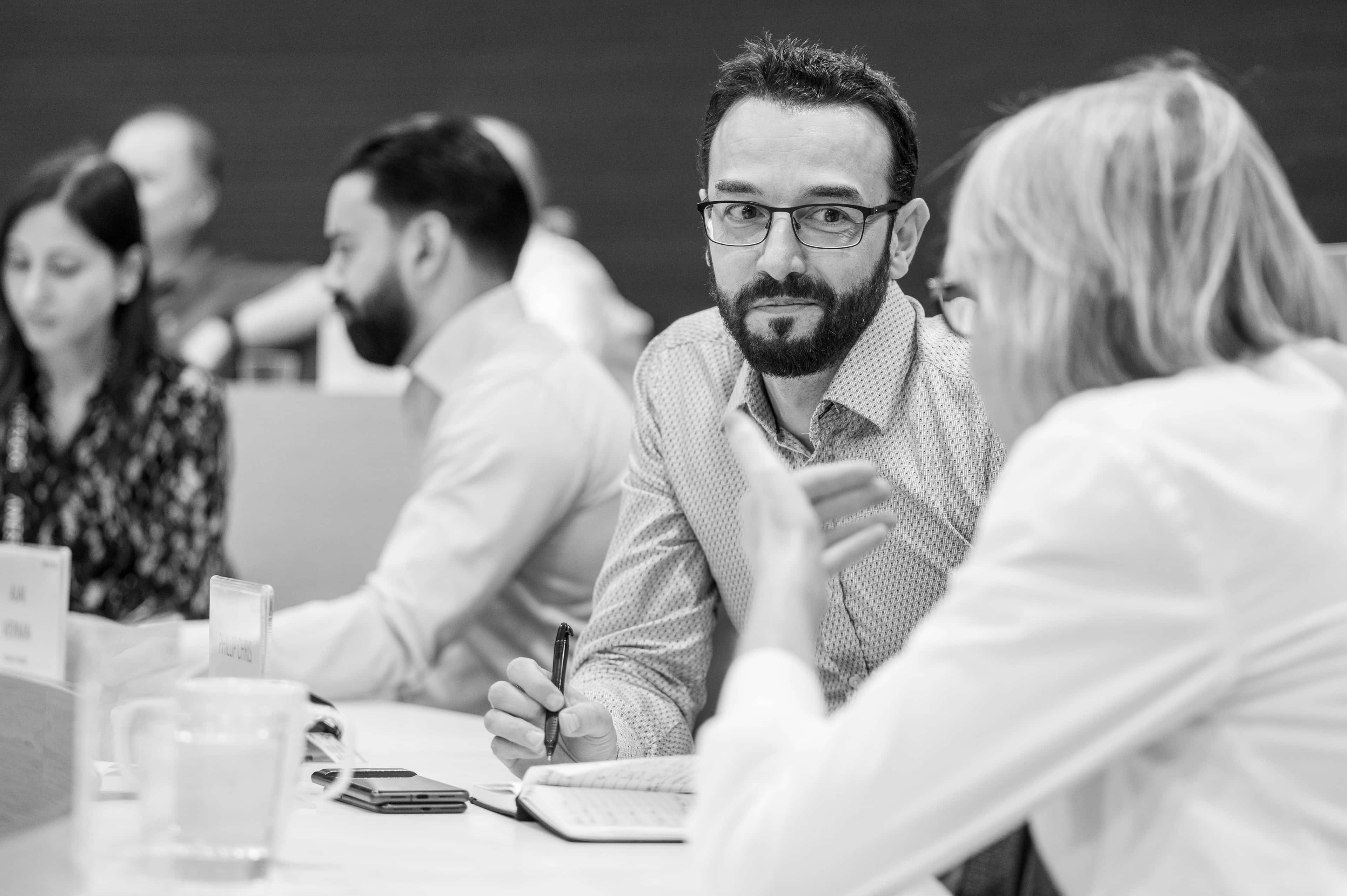 Bearded man with glasses listens attentively at a meeting, holding a pen and notebook while a colleague gestures beside him.