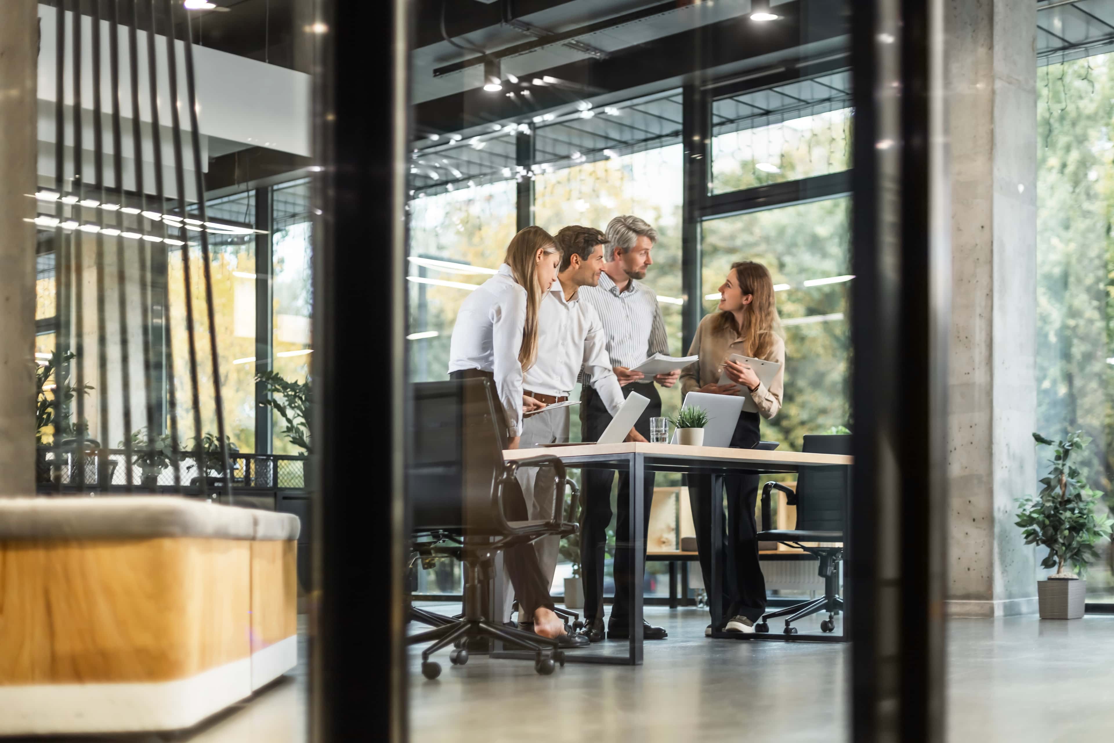 Four people in business attire stand around a table, discussing documents in a modern office with large windows and plants.