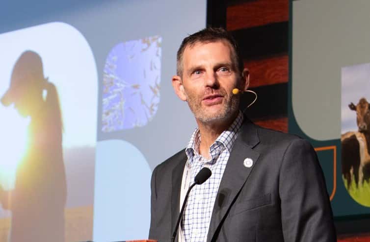 Man in a suit speaking at a conference with a microphone. Behind him, images of a silhouette, nature, and an animal are displayed on a screen.