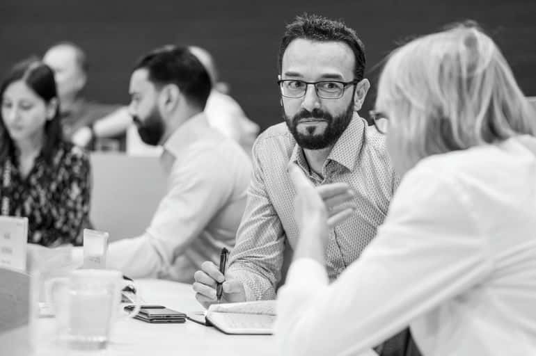 A man with glasses attentively listens at a conference table, holding a pen, with three other people engaged in conversation in the background.
