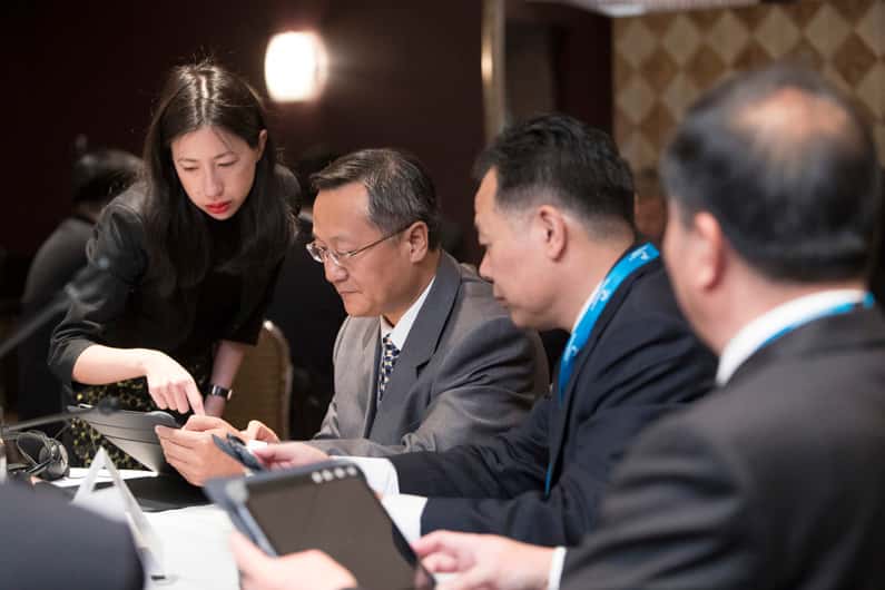 A woman in black shows something on a tablet to three men in suits seated at a conference table, engaging in discussion.