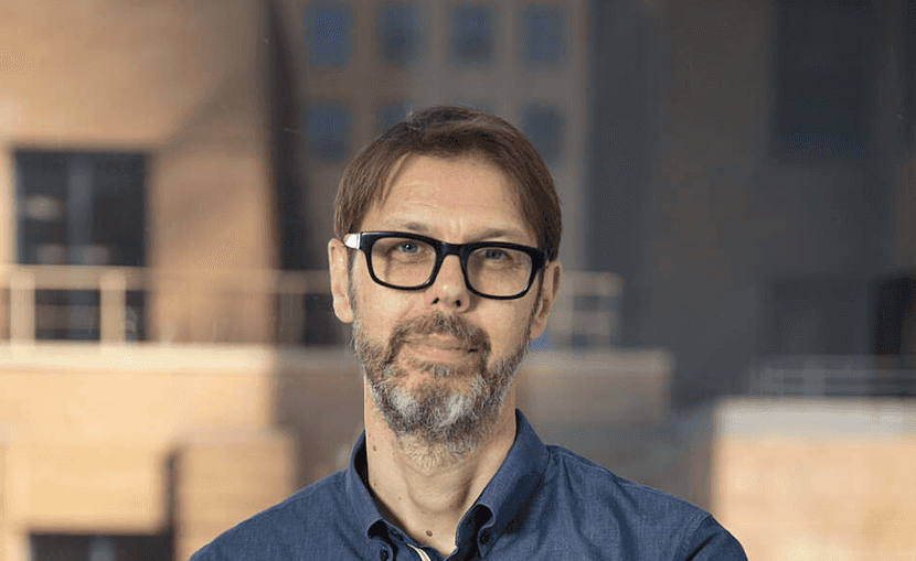 A man with dark glasses and a beard stands in front of an urban backdrop with blurred buildings, wearing a blue shirt.