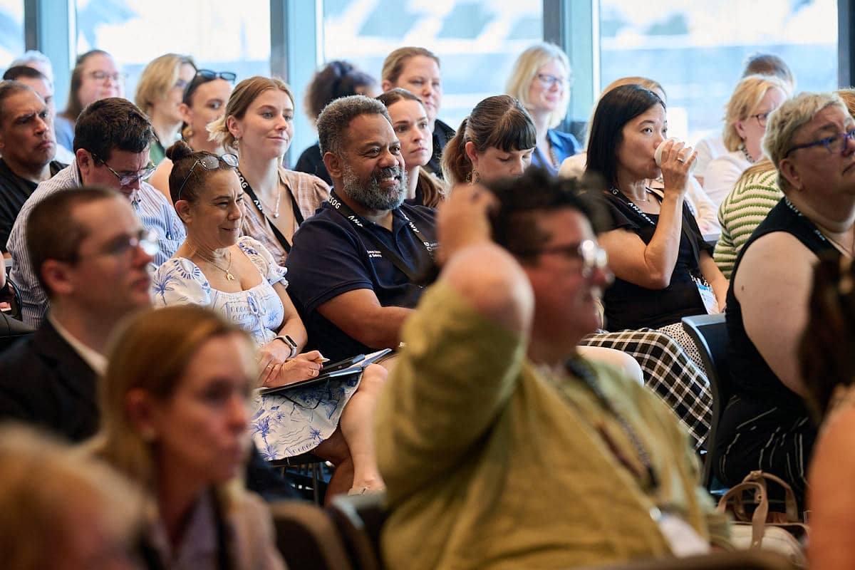 Conference audience of adults seated attentively; some smiling and taking notes, one woman sipping water.