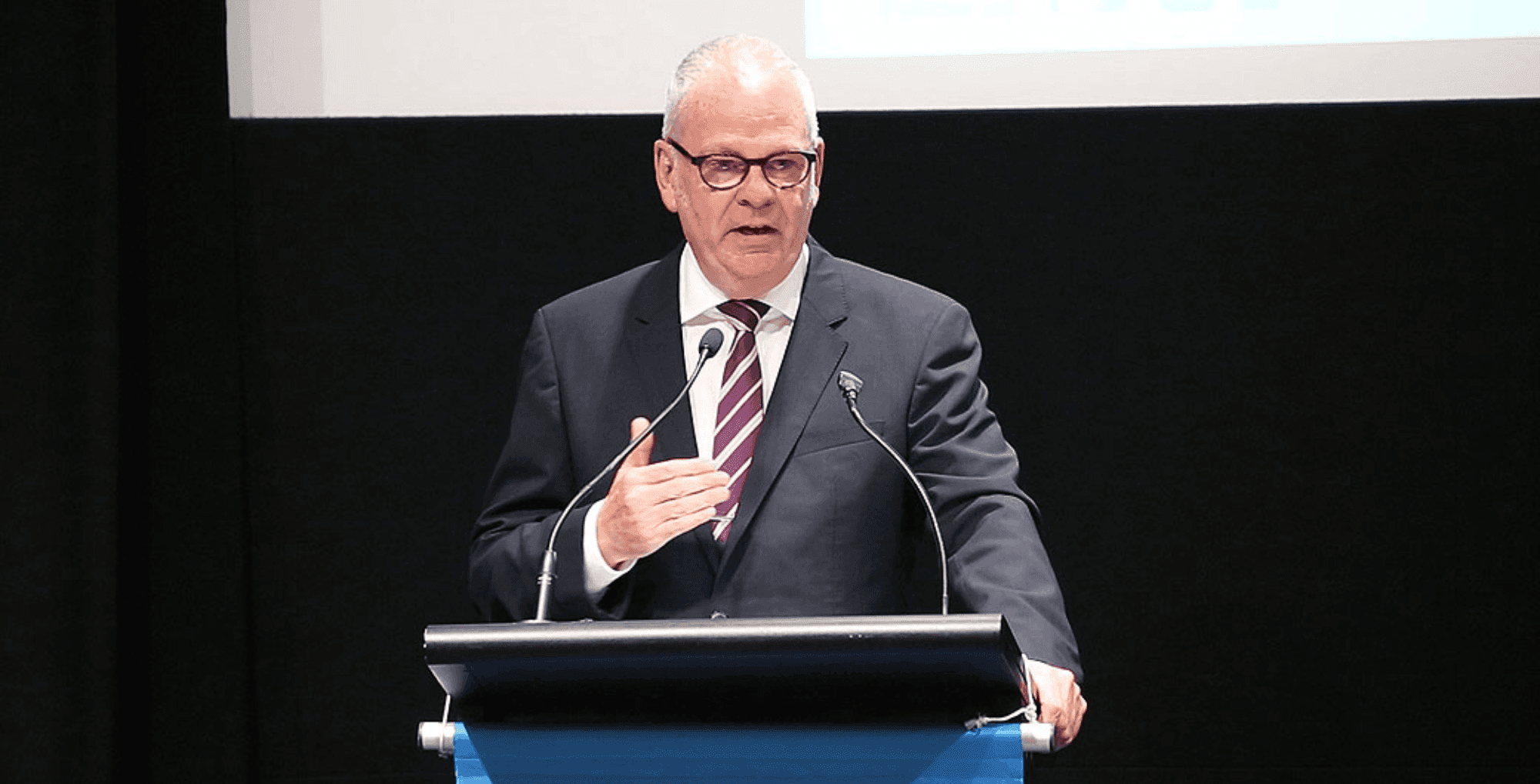 A man in a suit and tie speaks at a podium with two microphones, gesturing with his hand.