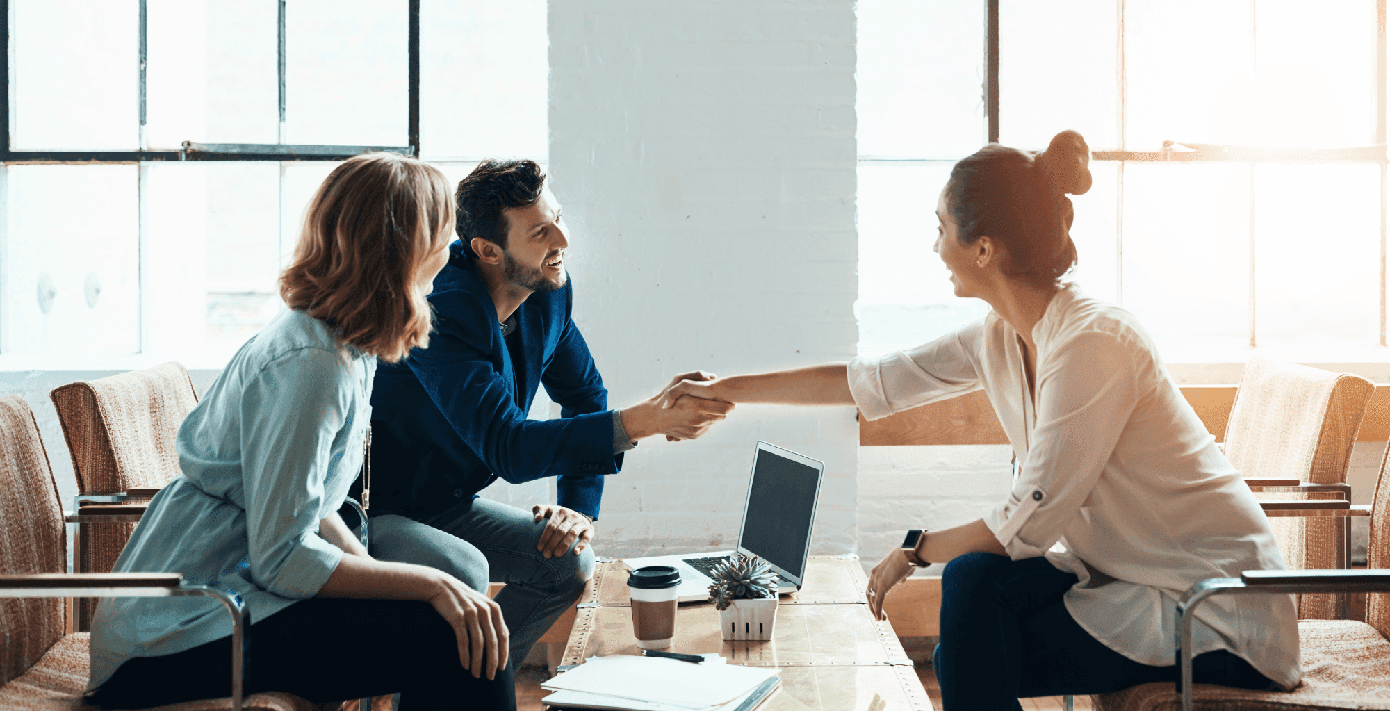 Three people are sitting in an office, two women and a man, smiling and shaking hands across a table with a laptop and coffee cup.