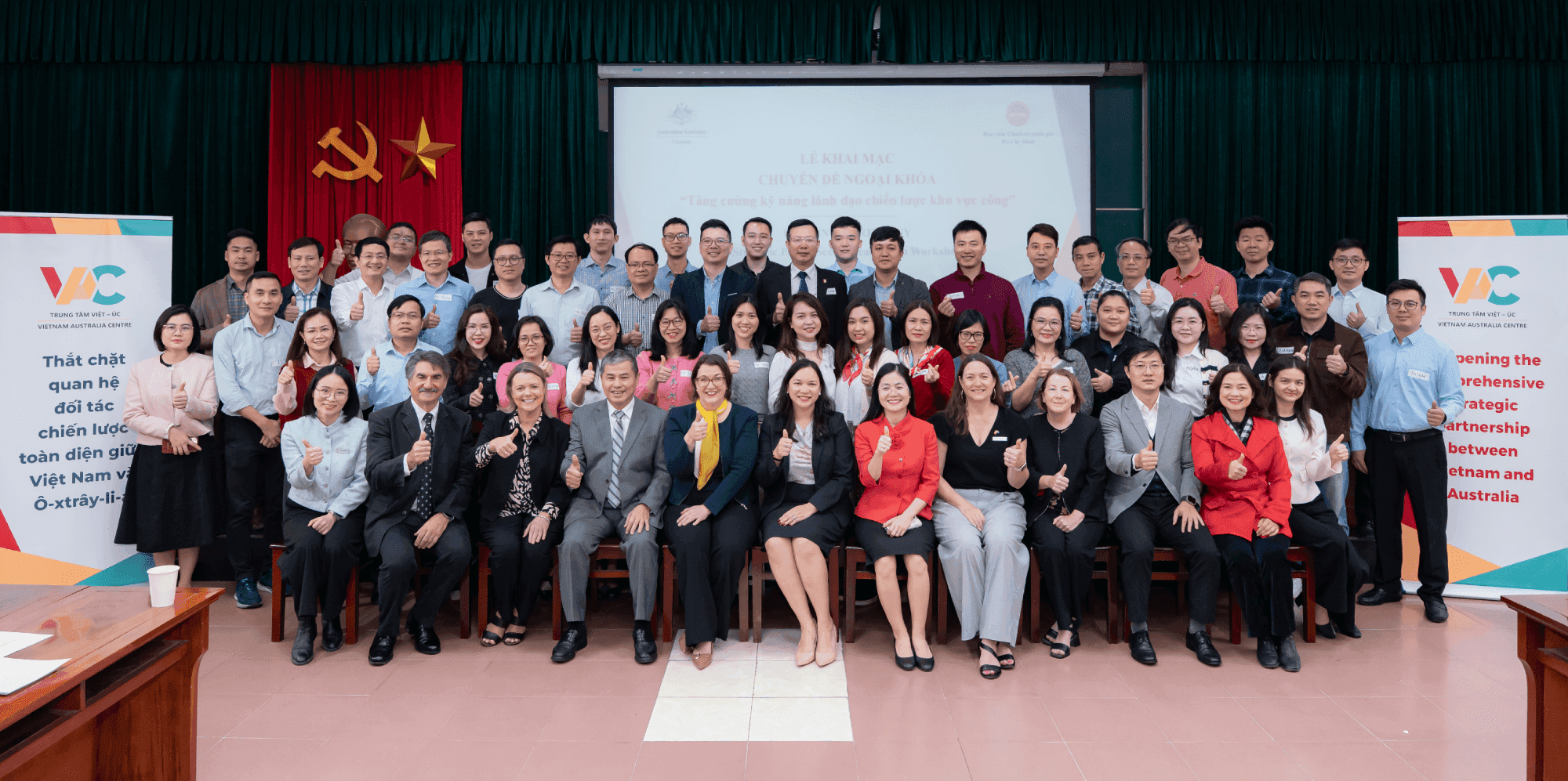 A large group of people posing indoors, with banners and a red curtain in the background. They are dressed in formal and semi-formal attire.