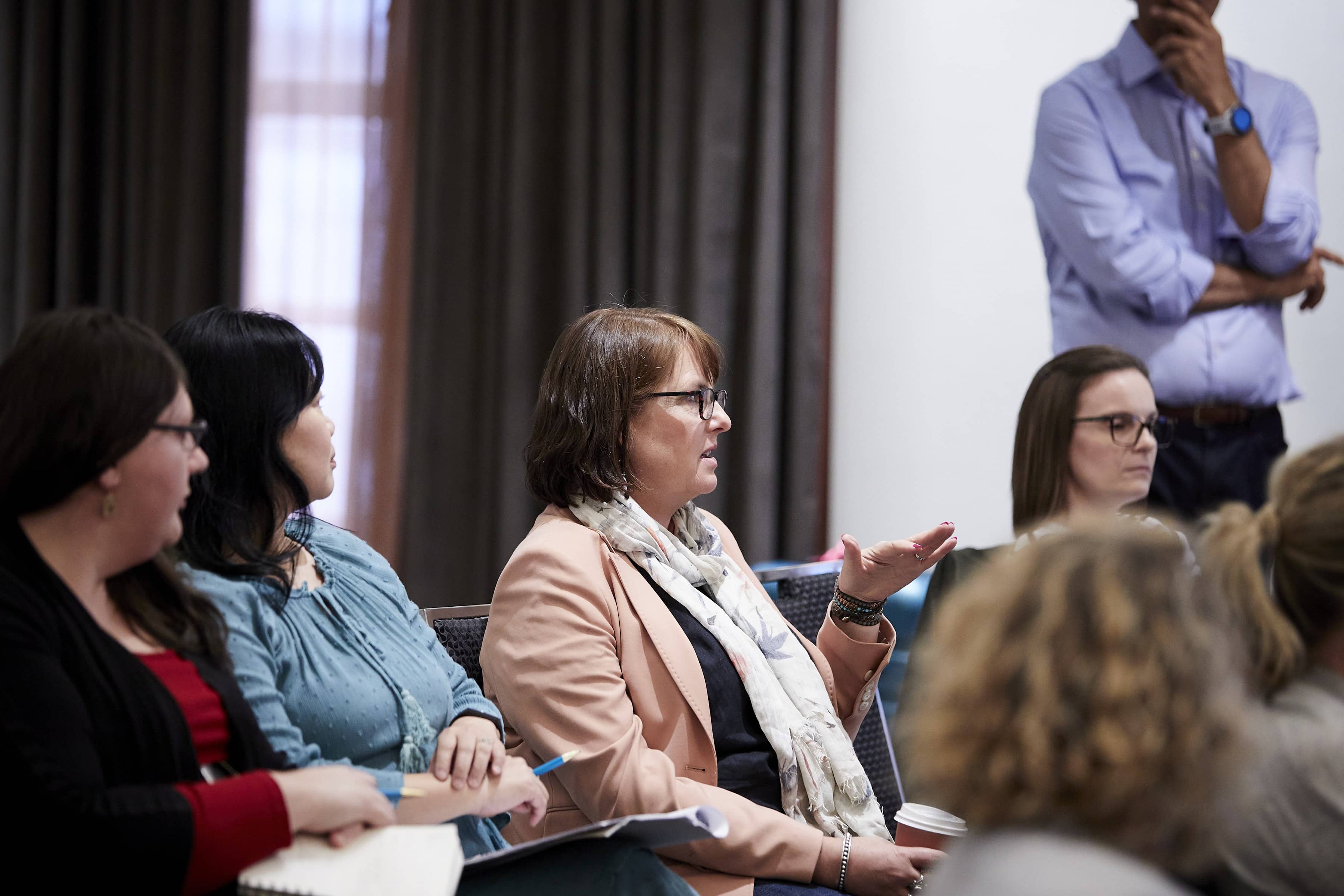 Woman gesturing while speaking at a meeting, seated among colleagues taking notes; a man stands listening in the background.