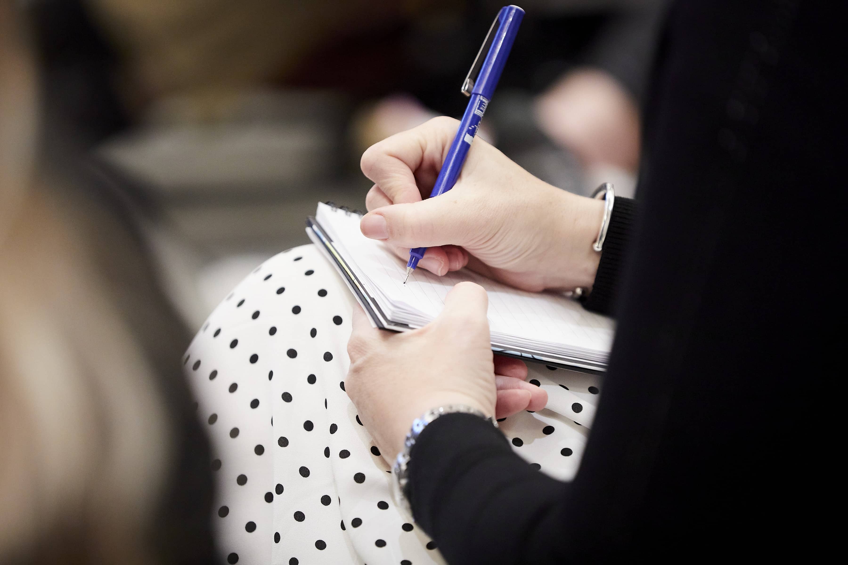 Close-up of hands writing with a blue pen in a spiral notebook resting on a white polka-dot skirt.