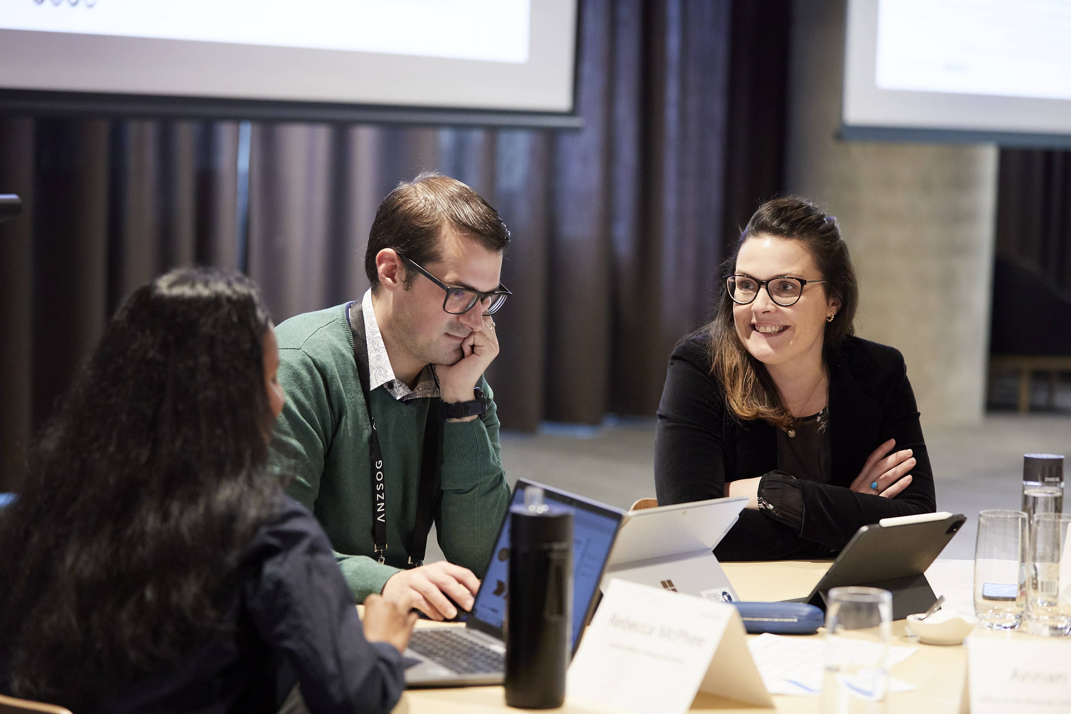 Three colleagues at a conference table collaborating over laptops; man in green sweater focused, woman with glasses smiling.