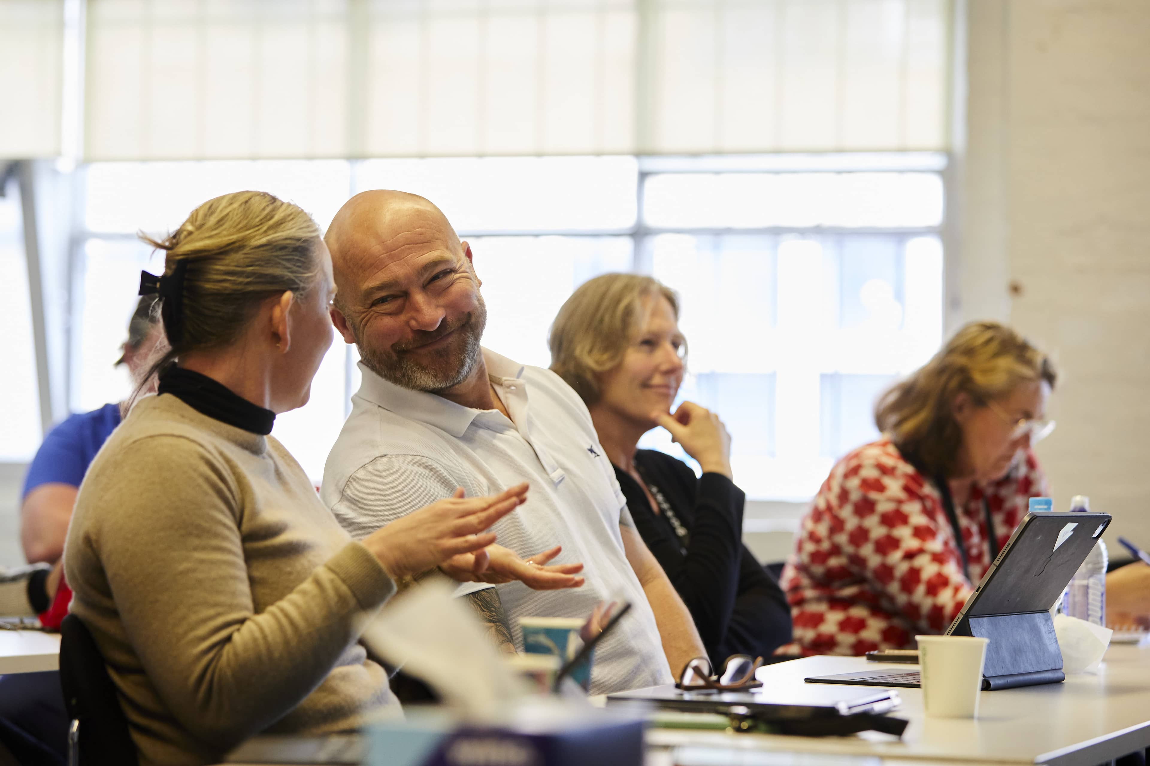 People seated in a bright classroom; smiling man leans toward a woman while others listen, write, and use a tablet.