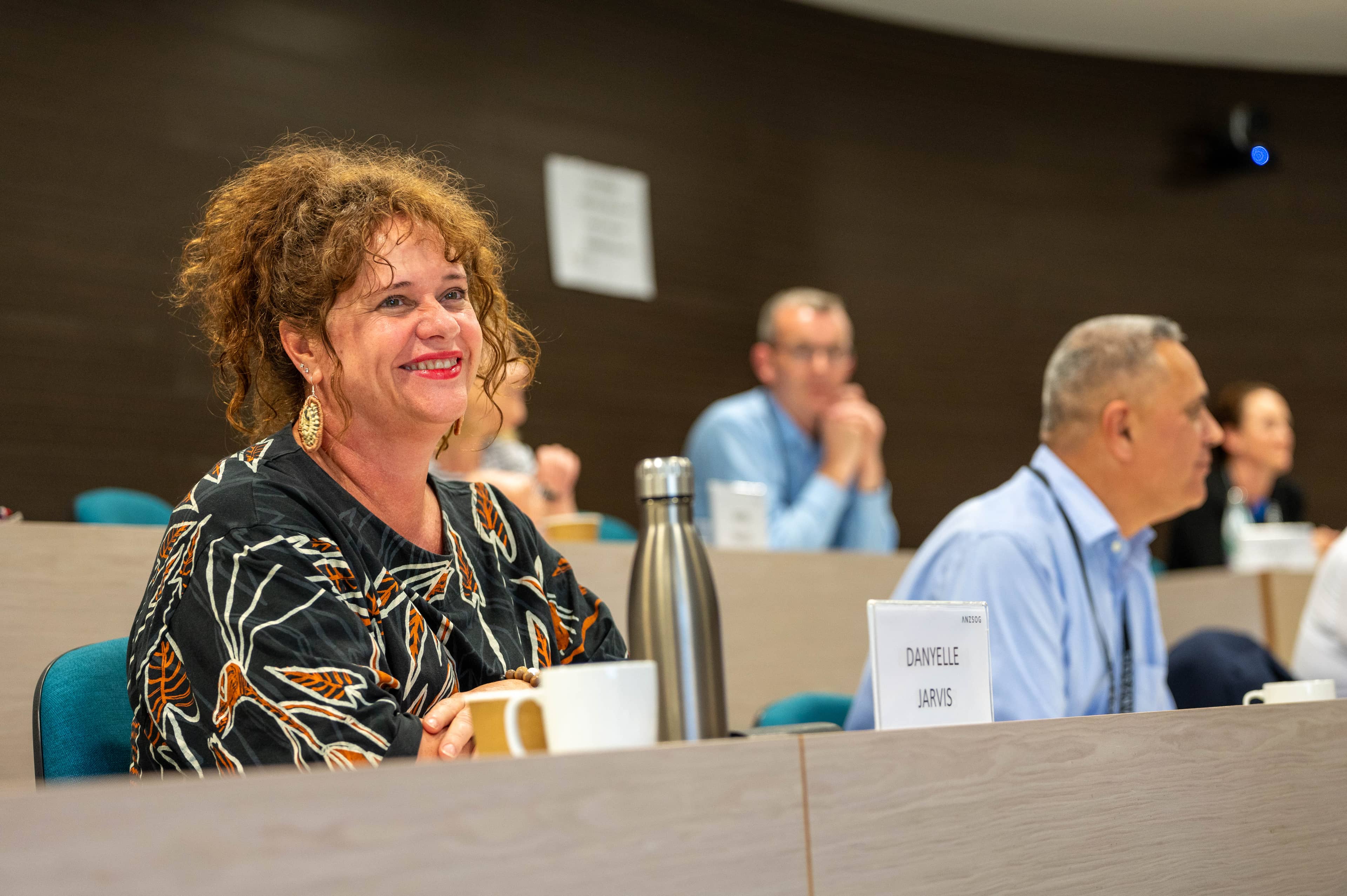 Smiling woman in a patterned blouse seated in a lecture hall, attentive, with a stainless water bottle and coffee mug on the desk.