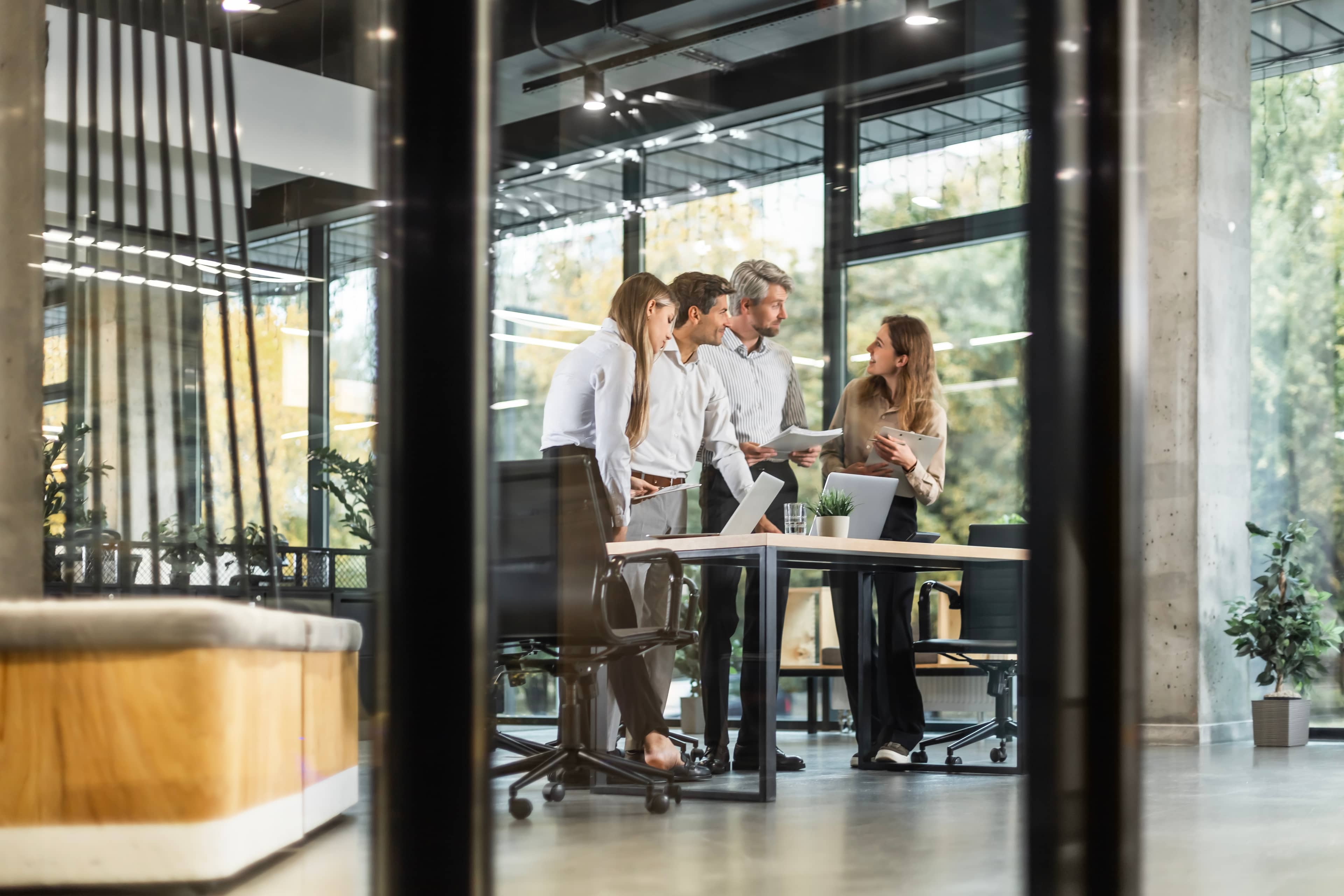 Four people in business attire stand around a table, discussing documents in a modern office with large windows and plants.