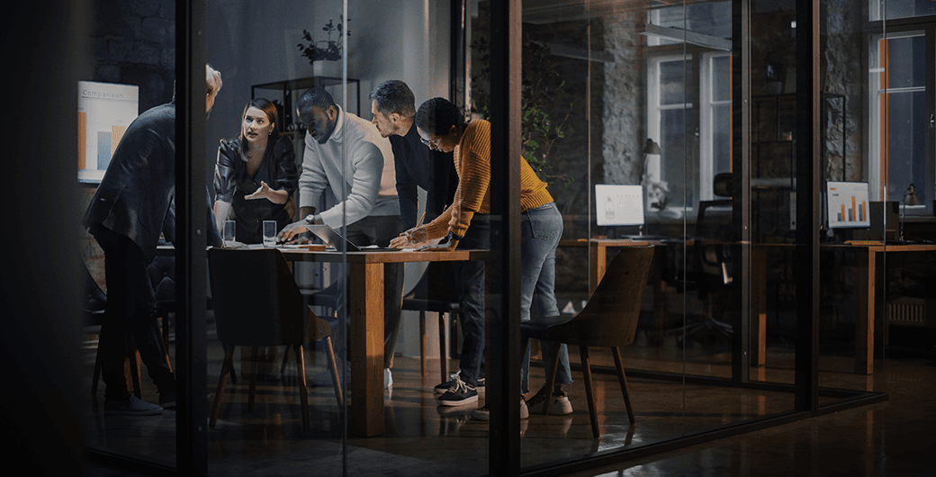 Group of professionals collaborating around a conference table in a modern glass-walled office at night, reviewing documents and laptops.