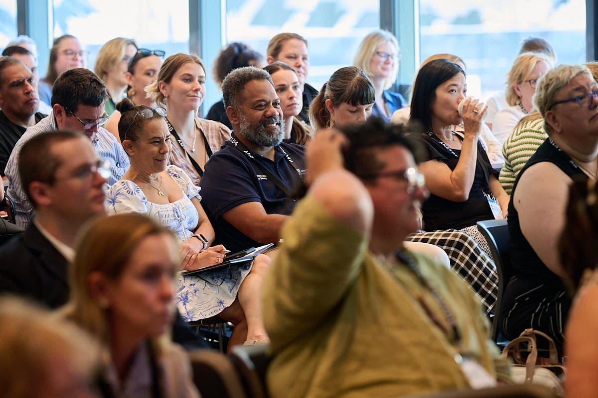 Conference audience of adults seated attentively; some smiling and taking notes, one woman sipping water.