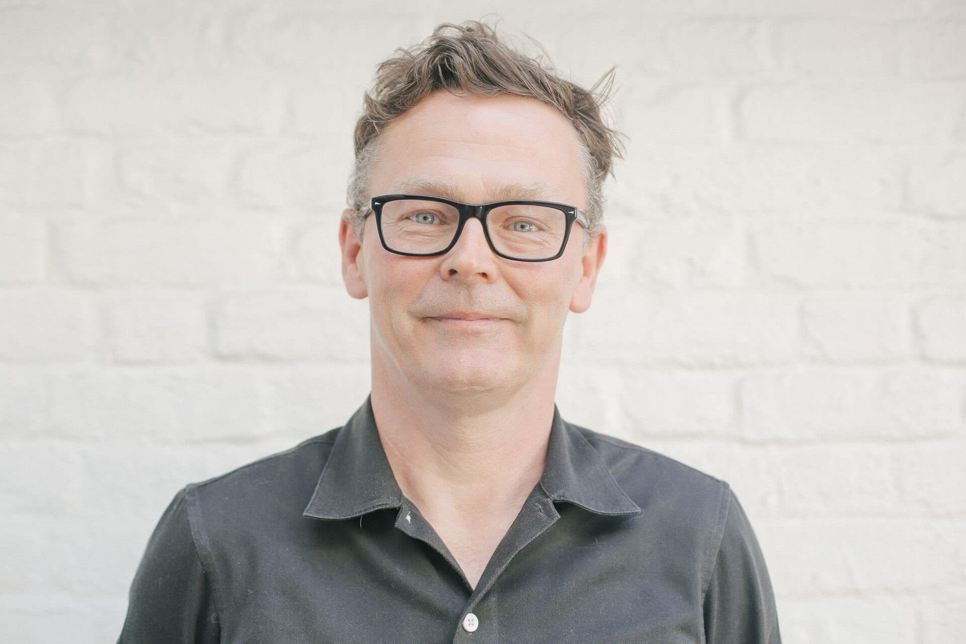 Man with glasses and short, wavy hair wearing a dark shirt, standing against a white brick wall.