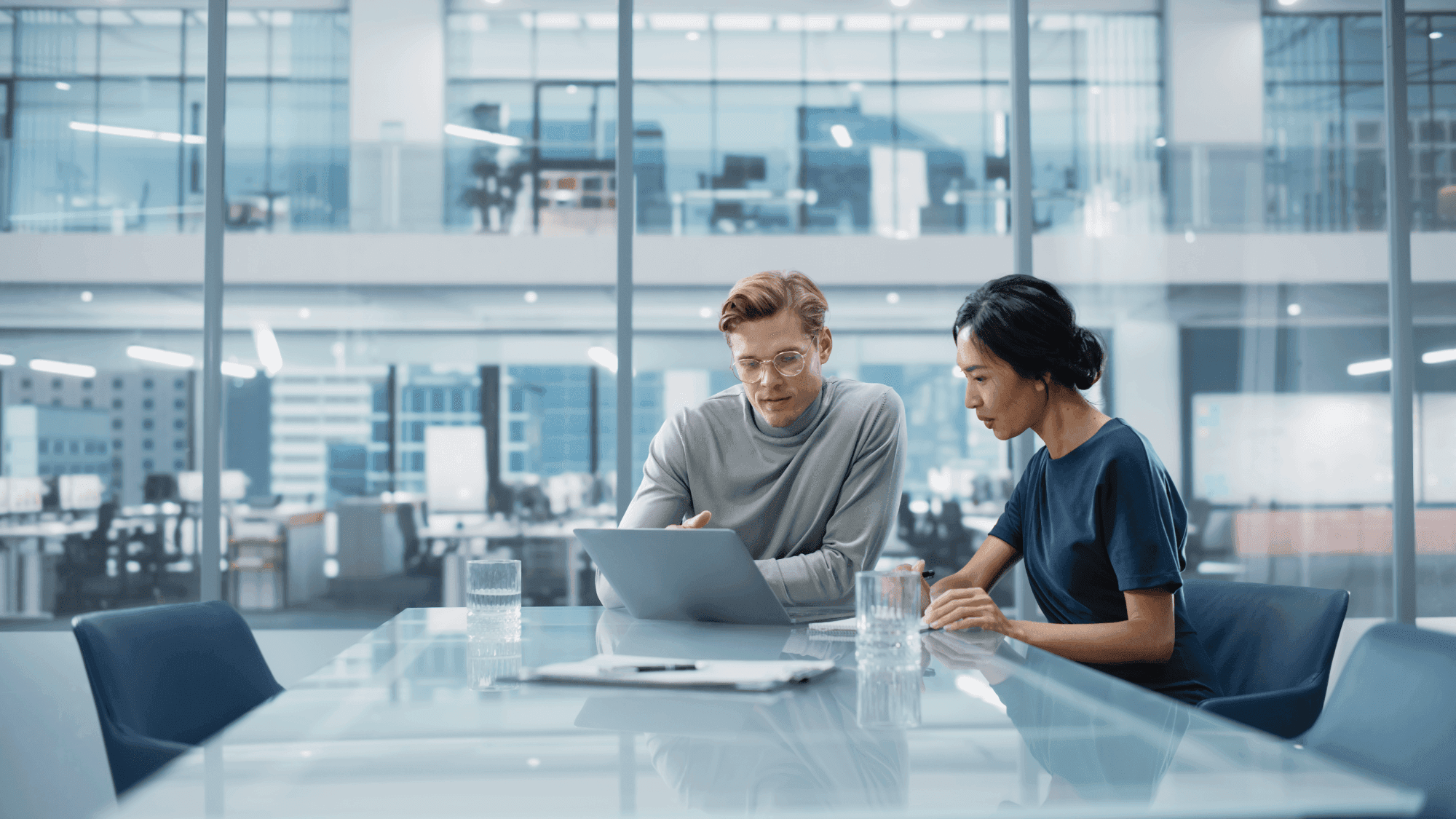Two people collaborate at a conference table with a laptop, in a modern office with glass walls and a city view.