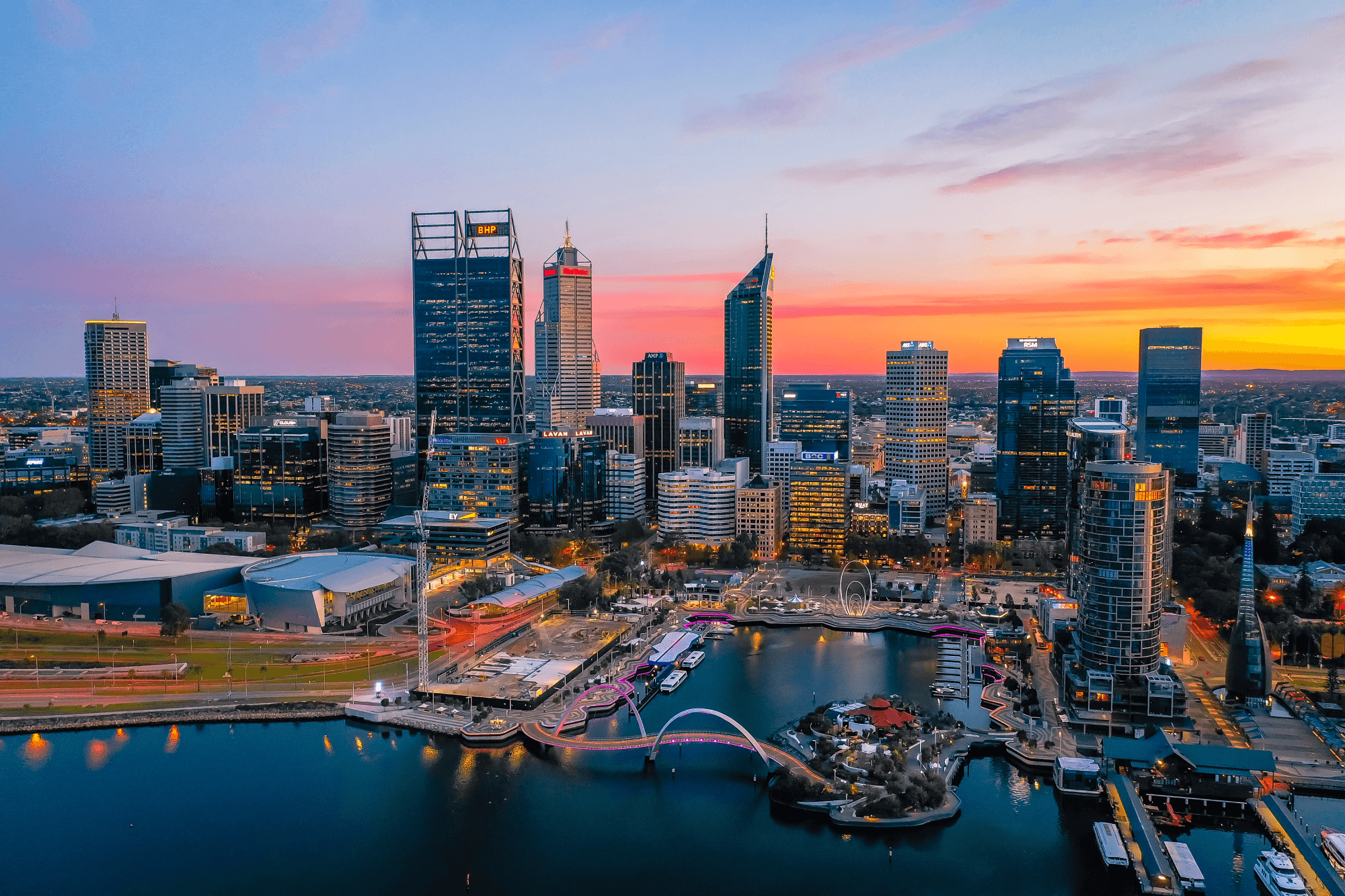 Aerial view of a city skyline at sunset, featuring tall skyscrapers, a waterfront, and a vibrant sky with orange and purple hues.